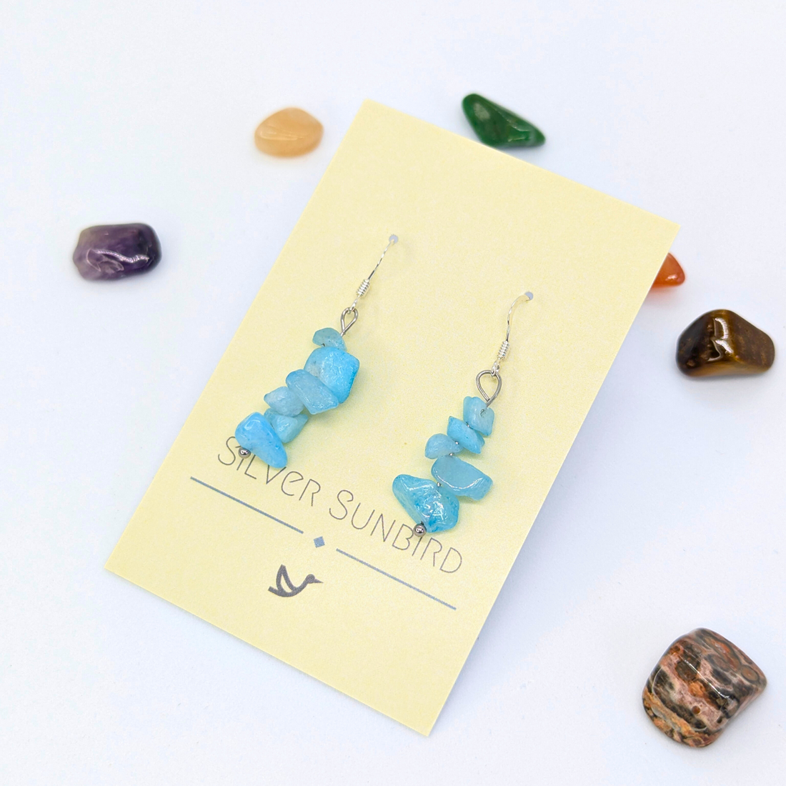 A pair of Aquamarine Gemstone Earrings by Silver Sunbird are displayed on a yellow brand card, surrounded by colorful polished stones, highlighting their handmade elegance against a white background.