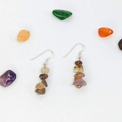 The Silver Sunbird Botswana Agate Gemstone Earrings feature stacked brown and beige gemstone chips, displayed on a white surface and surrounded by colorful polished stones.