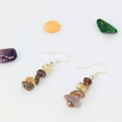 Silver Sunbird’s Botswana Agate Gemstone Earrings, featuring stacked multicolored stone beads, rest on a white surface surrounded by polished green, orange, and purple stones.