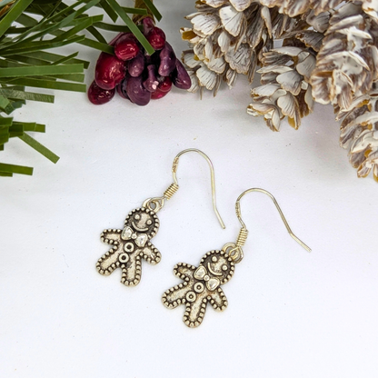 The Silver Sunbird Jolly Gingerbread Men Earrings feature a metallic finish and decorative details, shown on a white surface with pine branches, pine cones, and red berries.