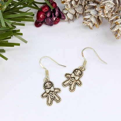 The Silver Sunbird Jolly Gingerbread Men Earrings, crafted from 925 sterling silver, are shown on a white surface with pine needles, red berries, and pinecones—ideal holiday jewelry for a festive accent.