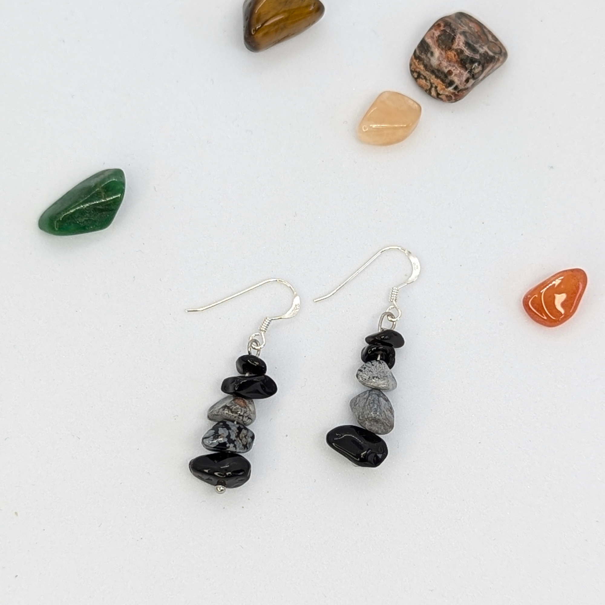 The Silver Sunbird Snowflake Obsidian Earrings feature stacked black and gray gemstone beads on silver hooks, displayed on a white surface surrounded by six small, colorful polished stones.