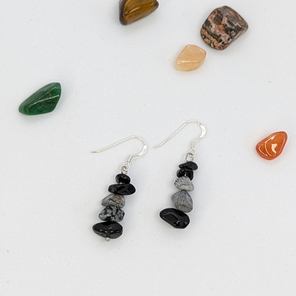 The Silver Sunbird Snowflake Obsidian Earrings feature stacked black and gray gemstone beads on silver hooks, displayed on a white surface surrounded by six small, colorful polished stones.
