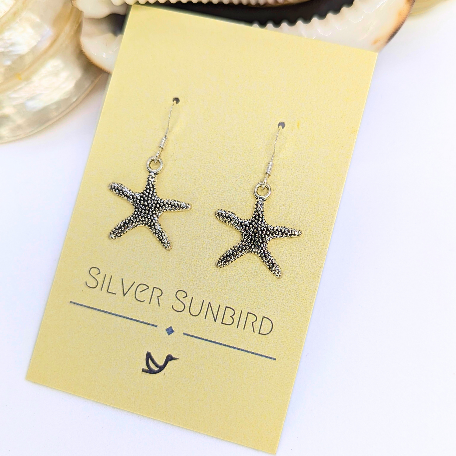 Detailed Silver Starfish Earrings by Silver Sunbird shown on a pale yellow card with a shell nearby—an ideal pick for lovers of unique boho jewellery.