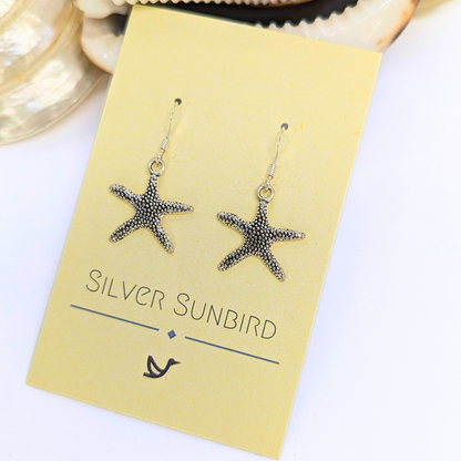 Detailed Silver Starfish Earrings by Silver Sunbird shown on a pale yellow card with a shell nearby—an ideal pick for lovers of unique boho jewellery.