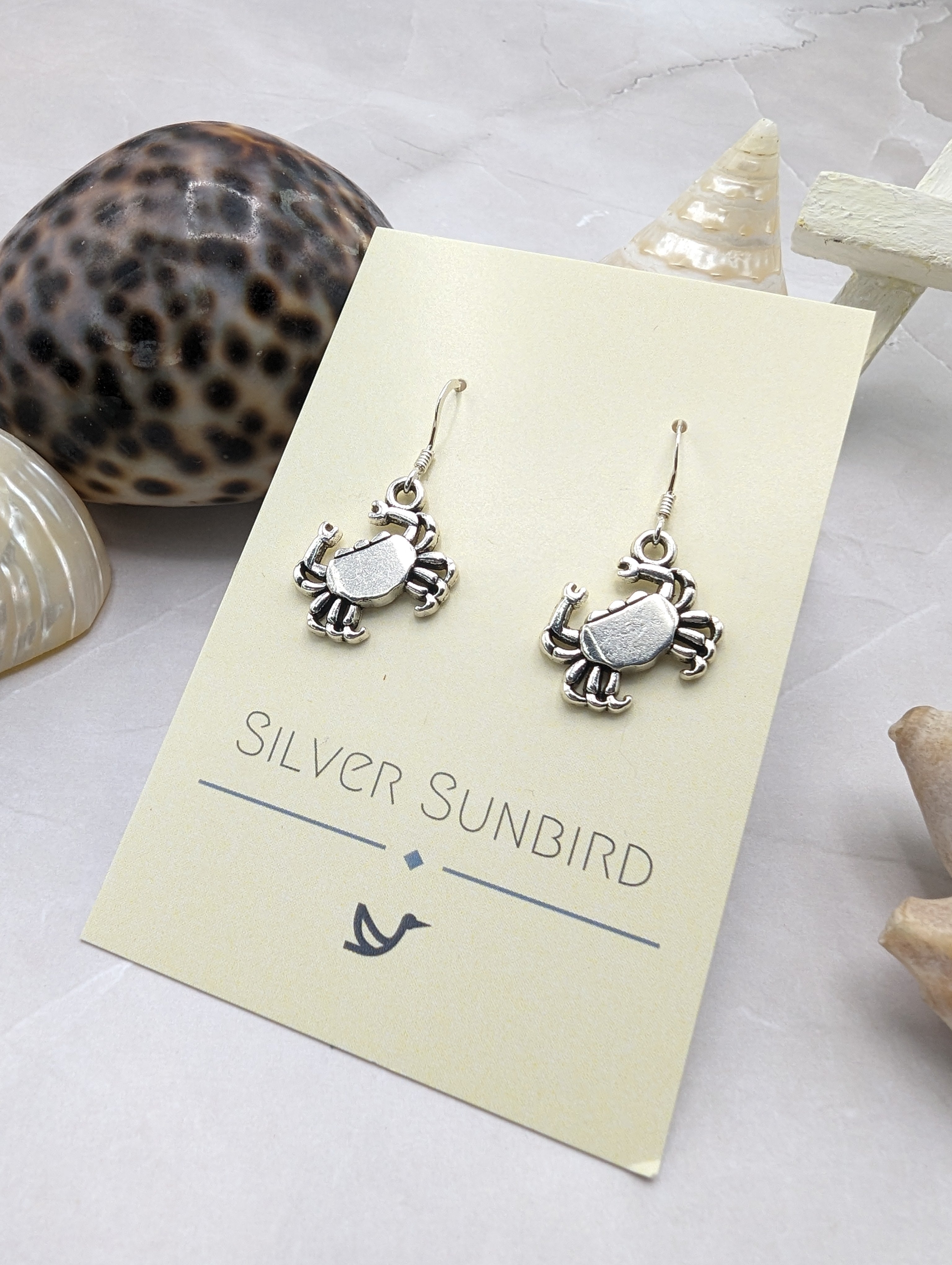 Displayed on a card with seashells, the &quot;Silver Sunbird&quot; Coastal Crab Earrings are sterling silver, crab-shaped, and hypoallergenic for comfort. These earrings bring seaside charm to any attire and offer a stylish nod to the coast.