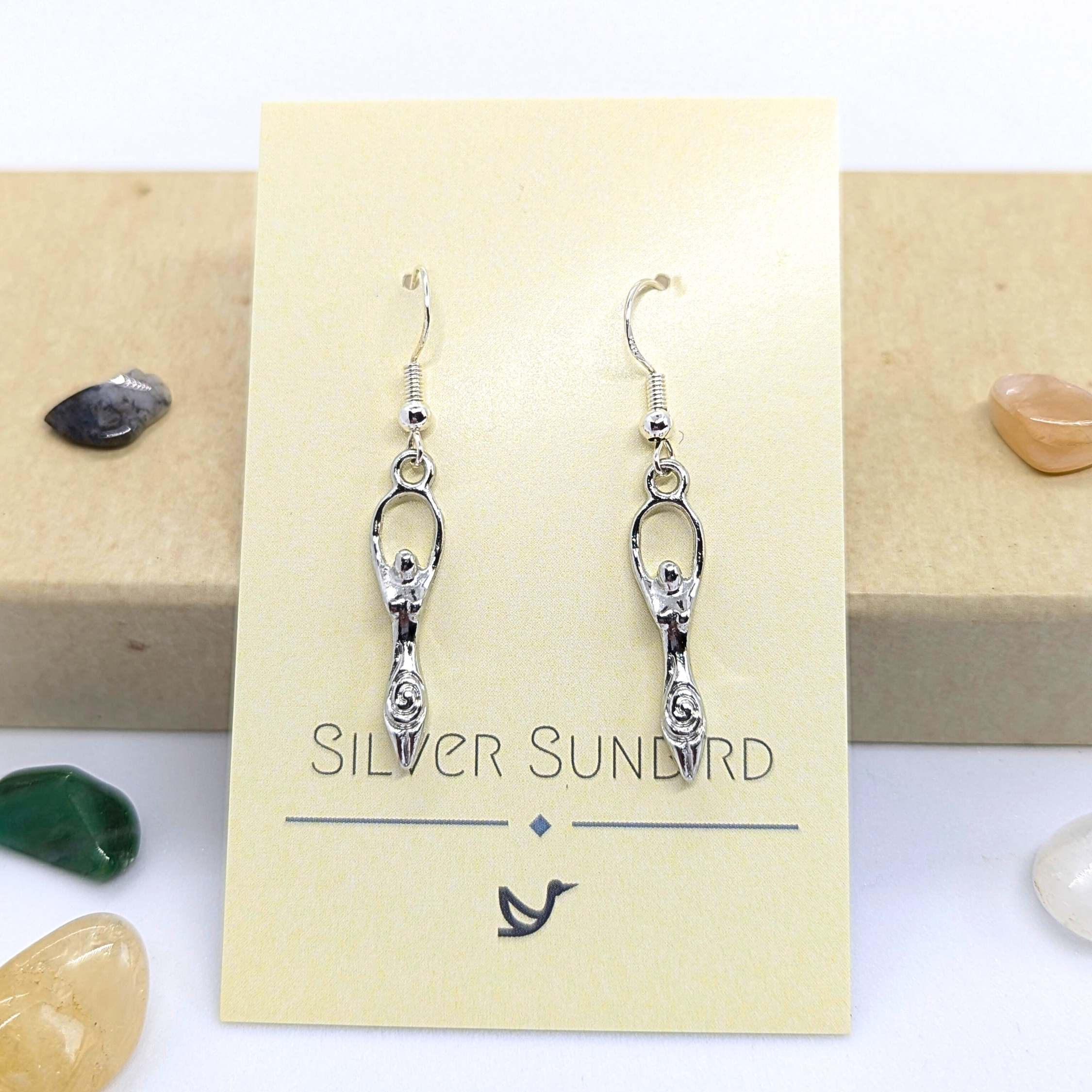 A pair of Fertility Goddess Dangle Earrings by Silver Sunbird is shown on a branded card, surrounded by polished green, yellow, brown, and black stones.