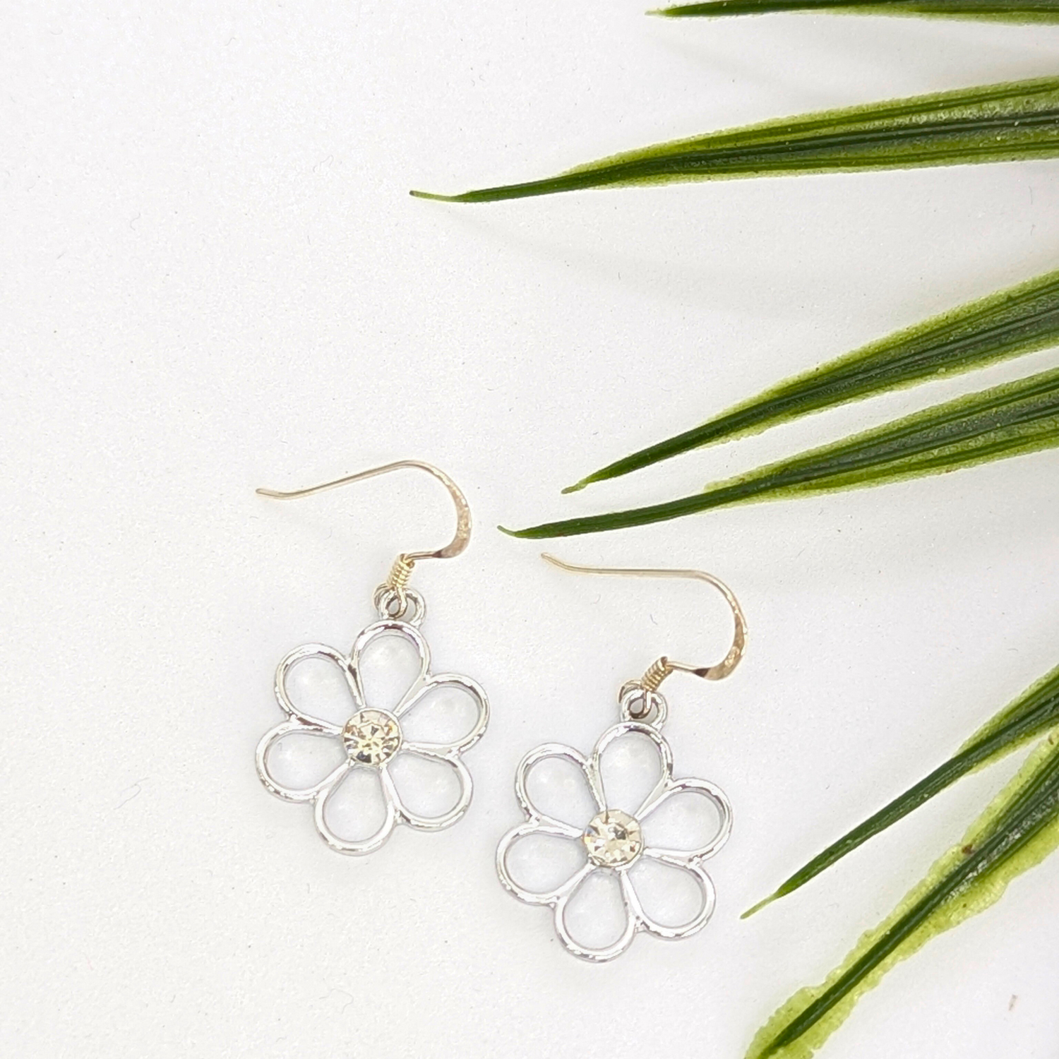The Sparkly Silver Daisy Earrings by Silver Sunbird, featuring a small central gem, rest on a white surface beside green spiky leaves, adding charm and elegance to these delicate floral accessories.