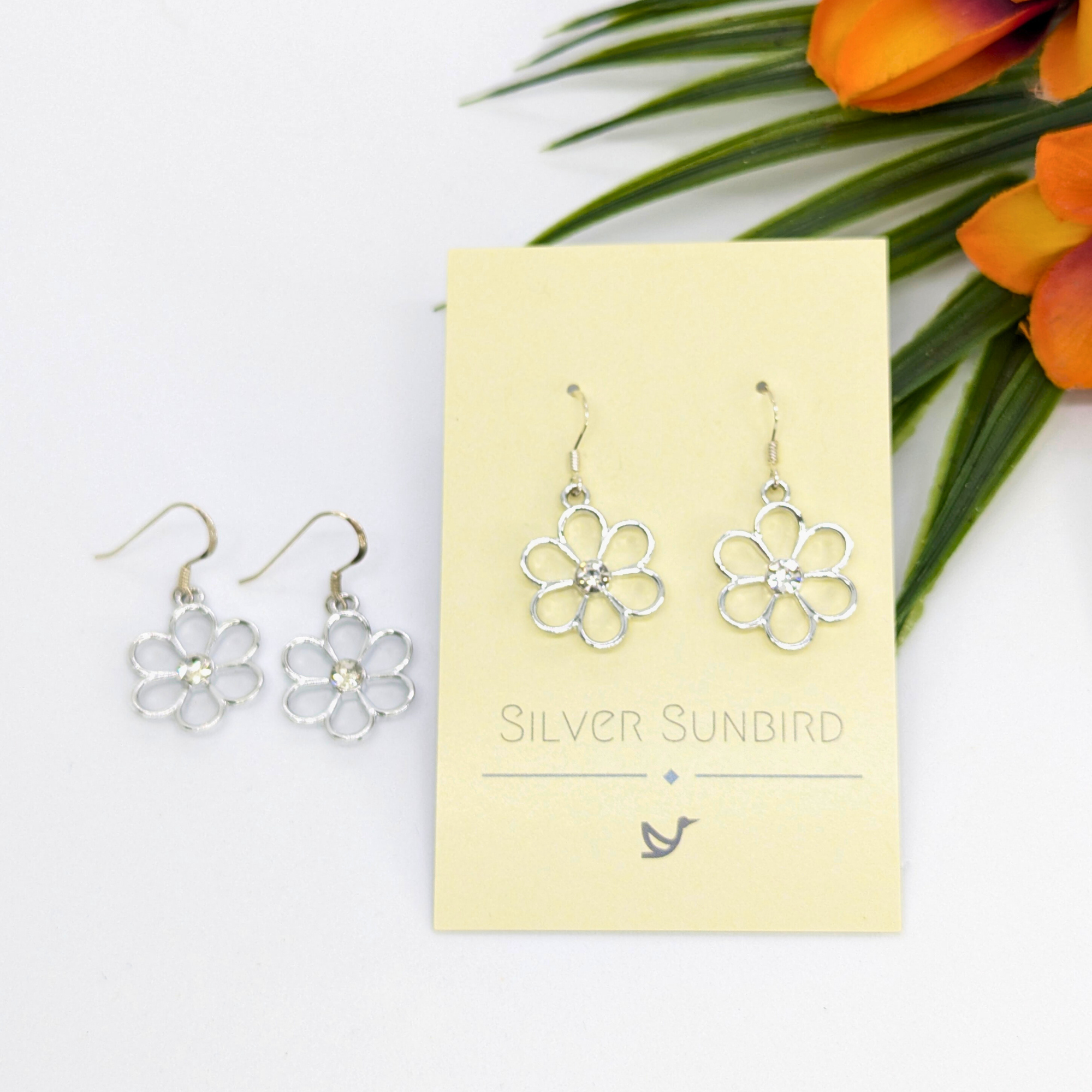 A pair of Sparkly Silver Daisy Earrings by Silver Sunbird—one set on a branded card, another beside it—with orange flowers and green leaves in the background.