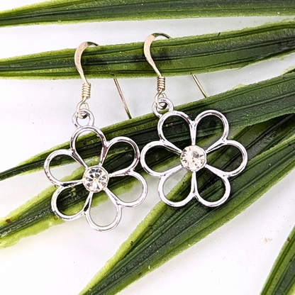 Sparkly Silver Daisy Earrings by Silver Sunbird feature clear gemstone centers and rest on green leaves against a white background. These enchanting floral earrings are crafted from sterling silver for an elegant touch.