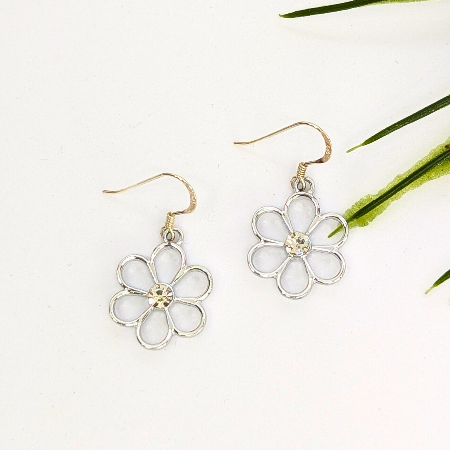 The Sparkly Silver Daisy Earrings by Silver Sunbird feature delicate open petals and a small central gem, showcased on a white background with green plant leaves in the corner.