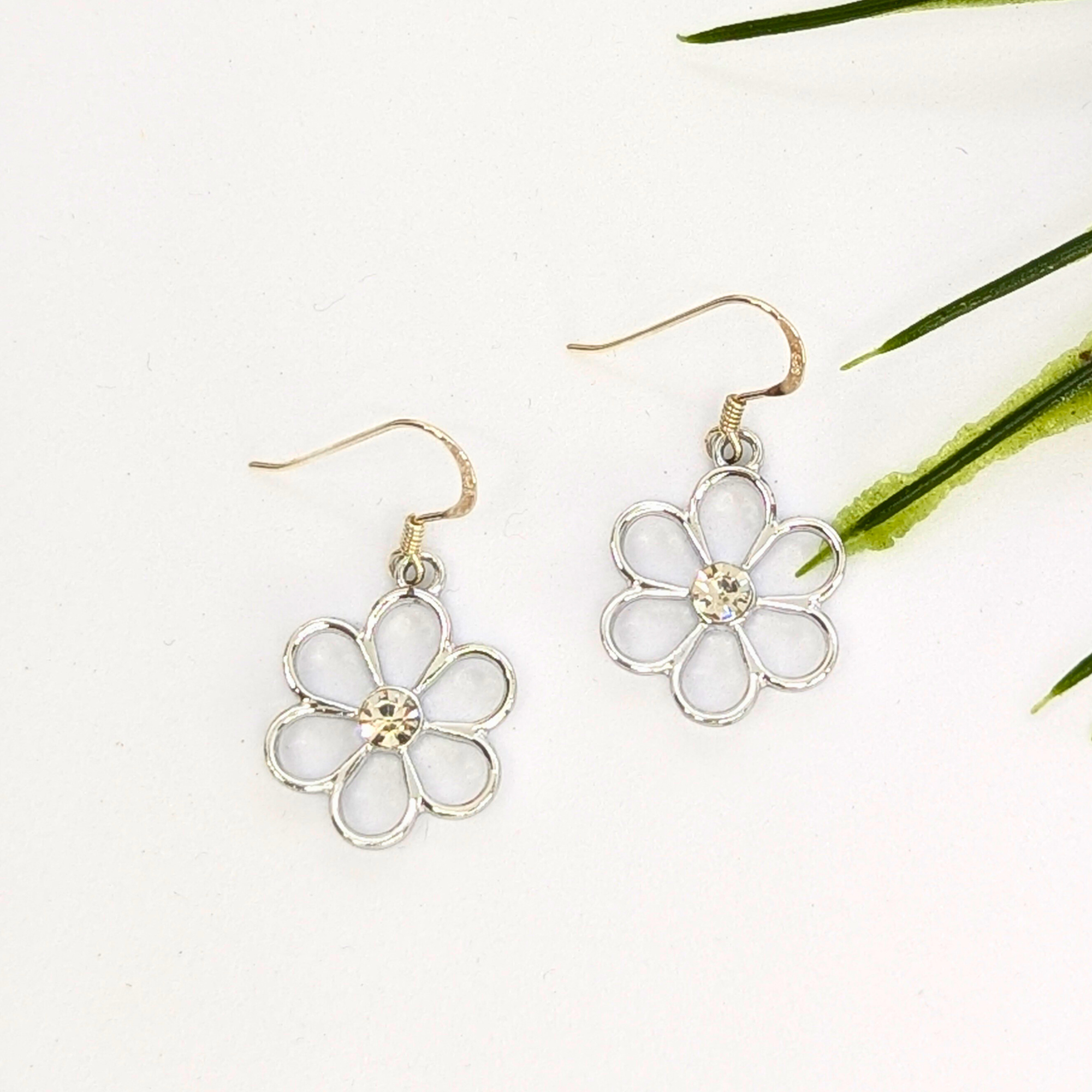 The Sparkly Silver Daisy Earrings by Silver Sunbird feature delicate open petals and a small central gem, showcased on a white background with green plant leaves in the corner.