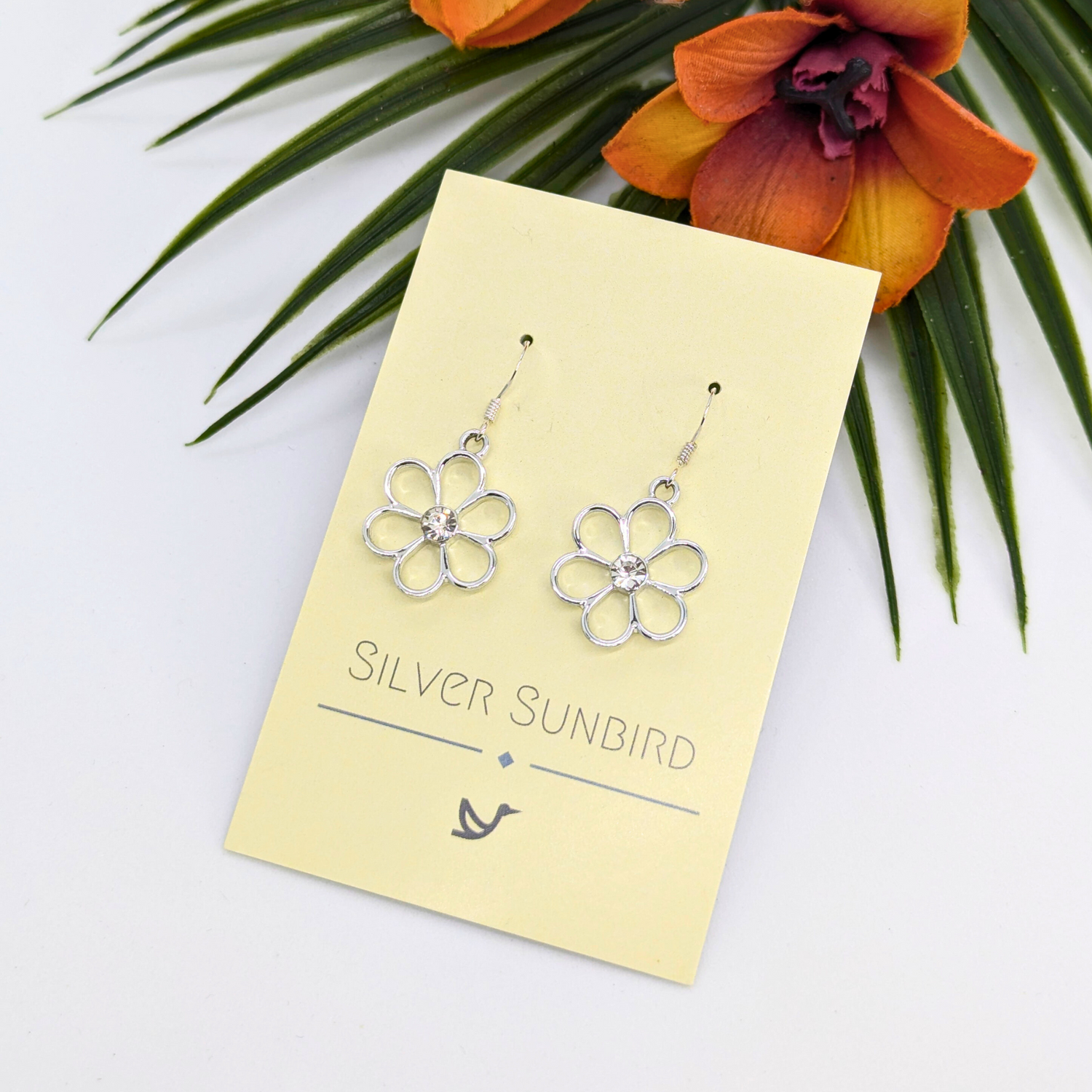 A pair of Sparkly Silver Daisy Earrings by Silver Sunbird, featuring a central gem, are displayed on a cream &quot;Silver Sunbird&quot; card with a tropical orange and purple flower and green leaves in the background.
