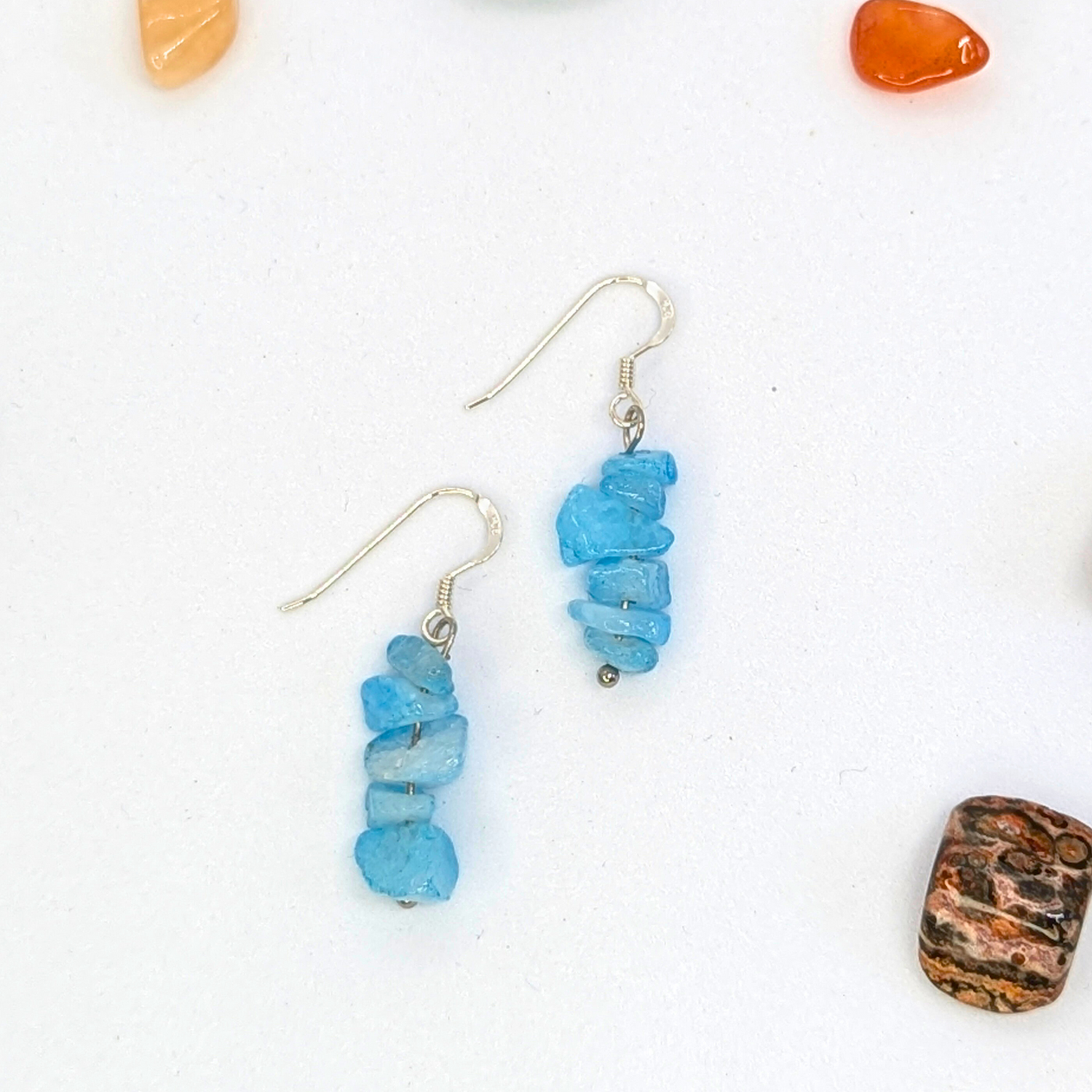 The Silver Sunbird Aquamarine Gemstone Earrings feature stacked irregular blue stones on sterling silver hypoallergenic hooks, displayed on a white surface with colorful stones scattered around.