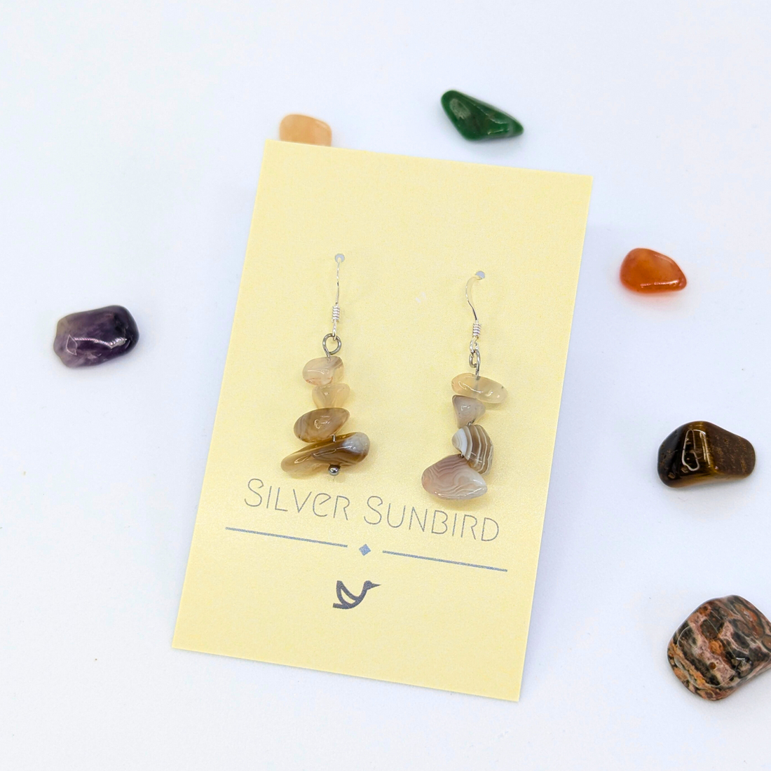 A pair of Botswana Agate Gemstone Earrings by Silver Sunbird is displayed on a cream card, surrounded by colorful polished stones against a white background.