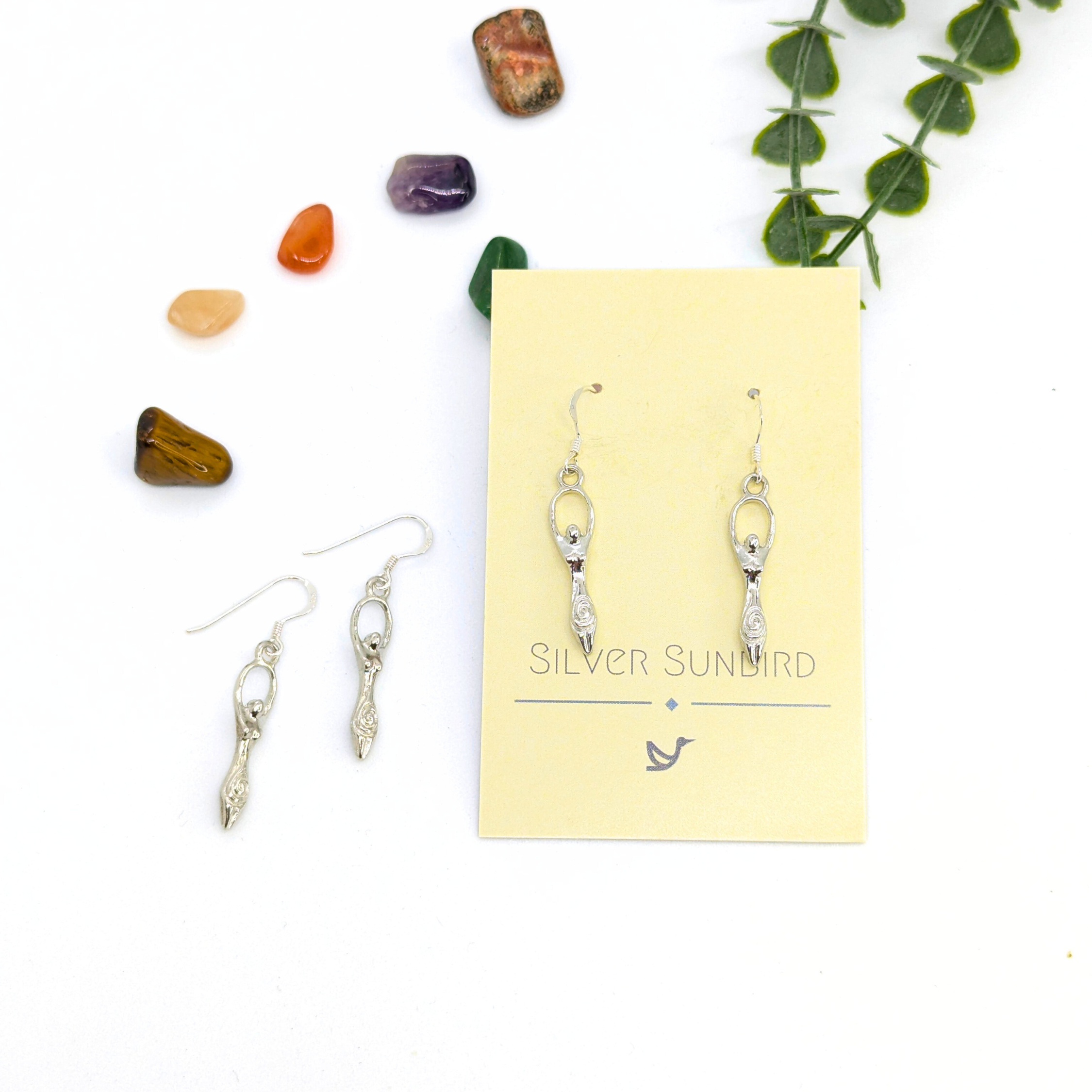 A yellow card labeled &quot;Silver Sunbird&quot; displays a pair of Fertility Goddess Dangle Earrings with abstract figures. To the left, another similar boho pair rests on a white surface among colorful crystals and green leaves.