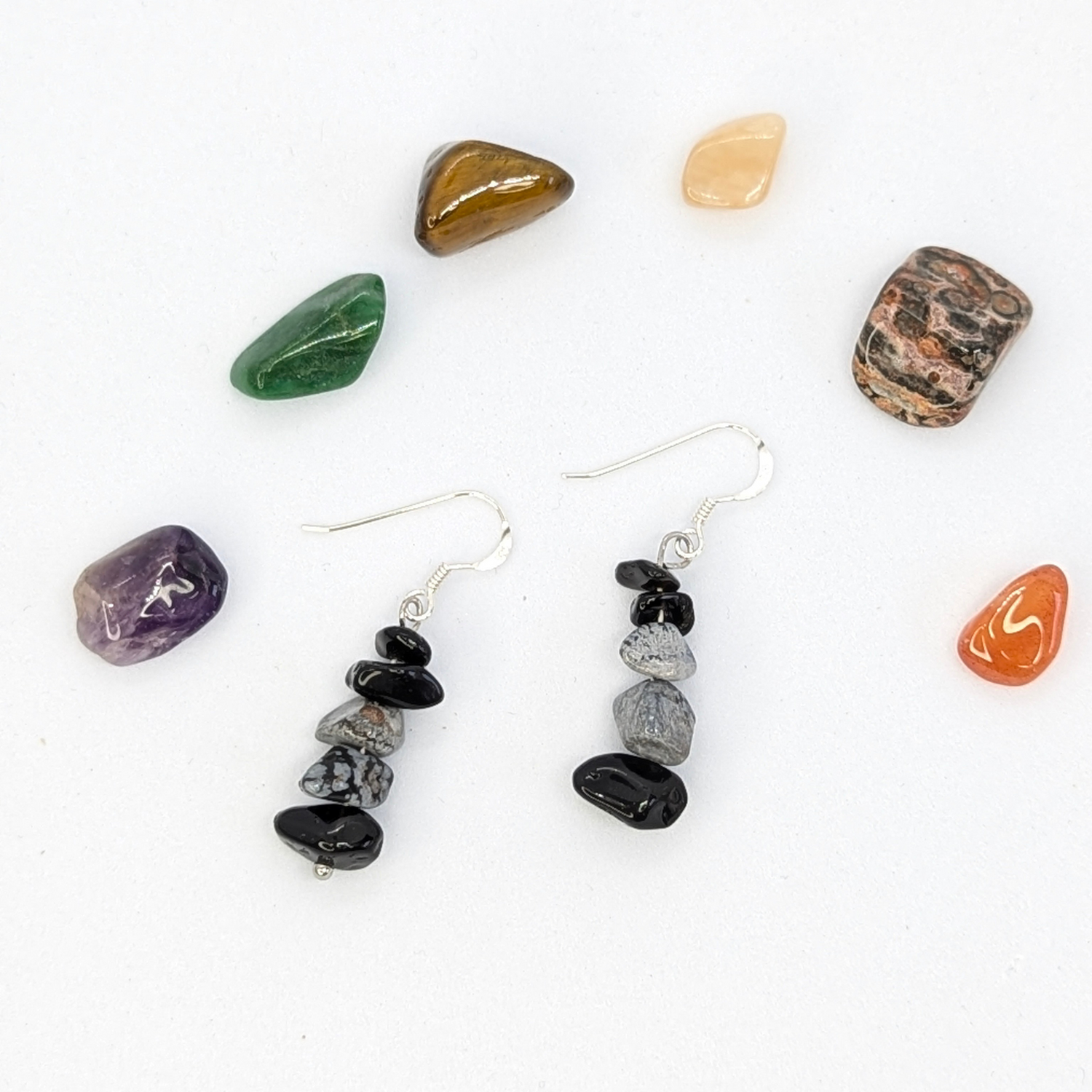 A pair of Silver Sunbird Snowflake Obsidian Earrings with stacked black and gray stones rests on a white surface, encircled by seven colorful, polished gemstones of assorted shapes.