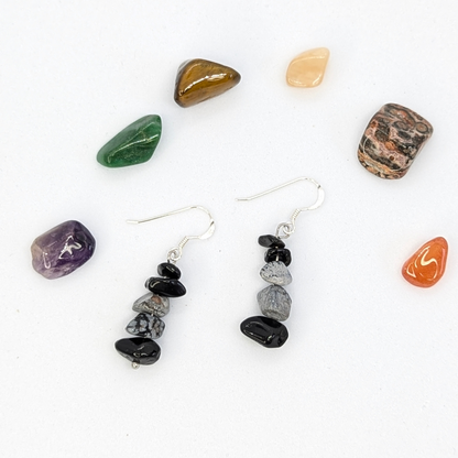 A pair of Silver Sunbird Snowflake Obsidian Earrings with stacked black and gray stones rests on a white surface, encircled by seven colorful, polished gemstones of assorted shapes.