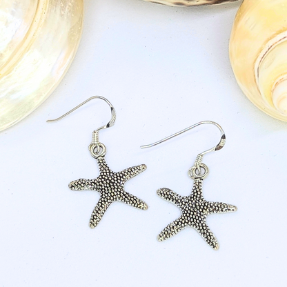 The Detailed Silver Starfish Earrings by Silver Sunbird are shown on a white surface with light seashells in the background—perfect boho jewellery to enhance your style.