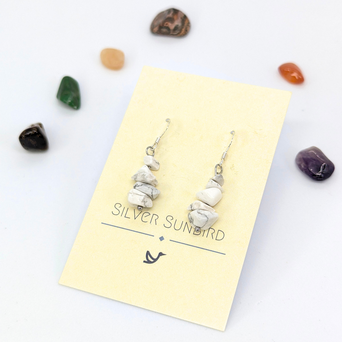 A pair of Silver Sunbird White Howlite Gemstone Earrings with silver hooks is shown on a cream card labeled &quot;SILVER SUNBIRD,&quot; surrounded by small polished stones against a white background.