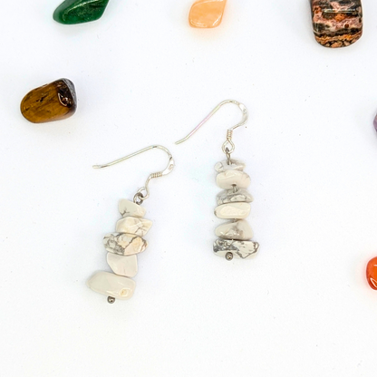 The Silver Sunbird White Howlite Gemstone Earrings feature stacked white and gray marbled stones on silver hooks, elegantly displayed with colorful polished stones.