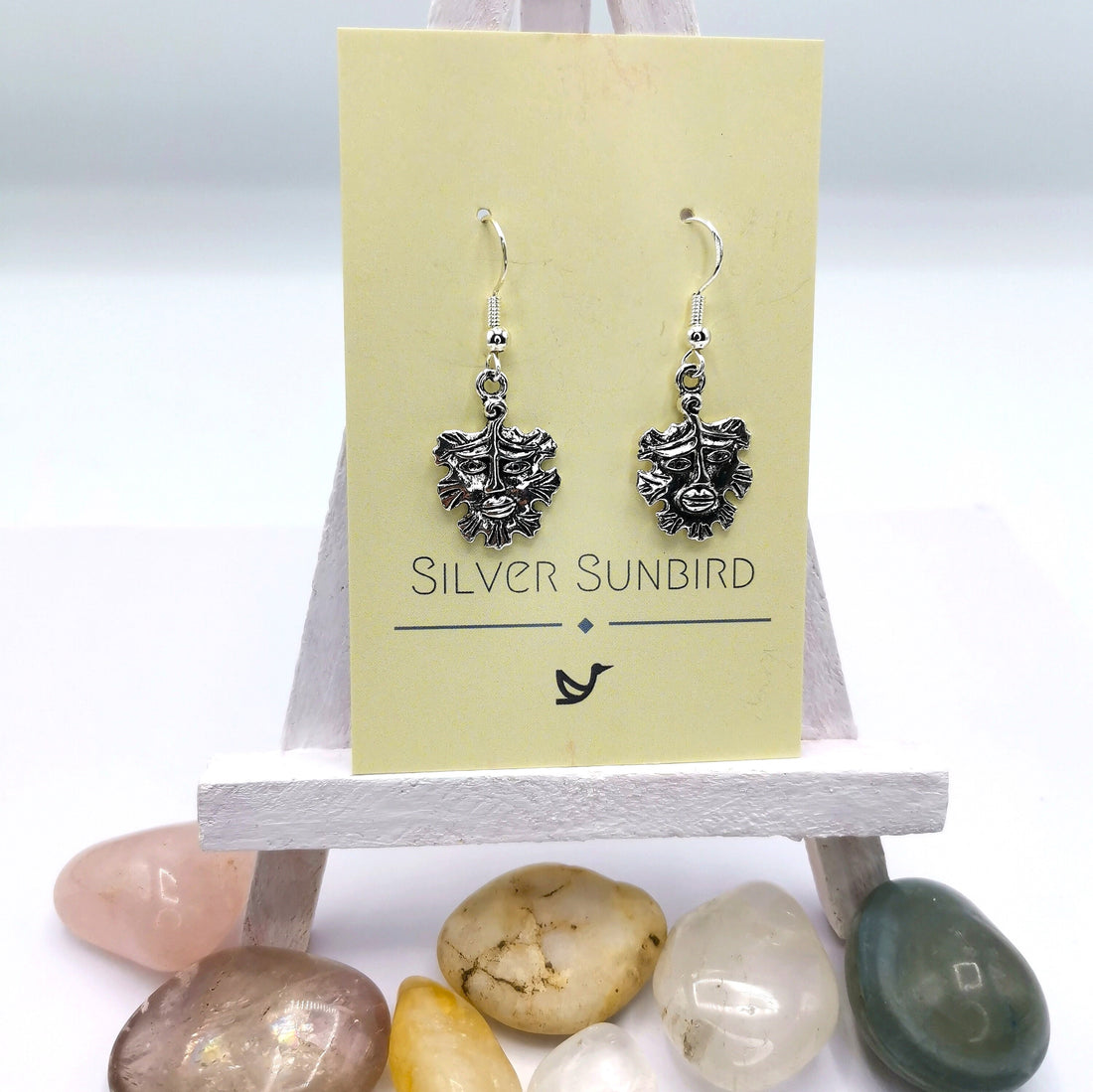 A pair of 925 sterling silver earrings with tribal sun and bird designs, presented on a card labeled "Gentle Green Man Silver Earrings" by Silver Sunbird. Displayed on a miniature easel with colorful stones, these earrings exude the charm of pagan jewelry.