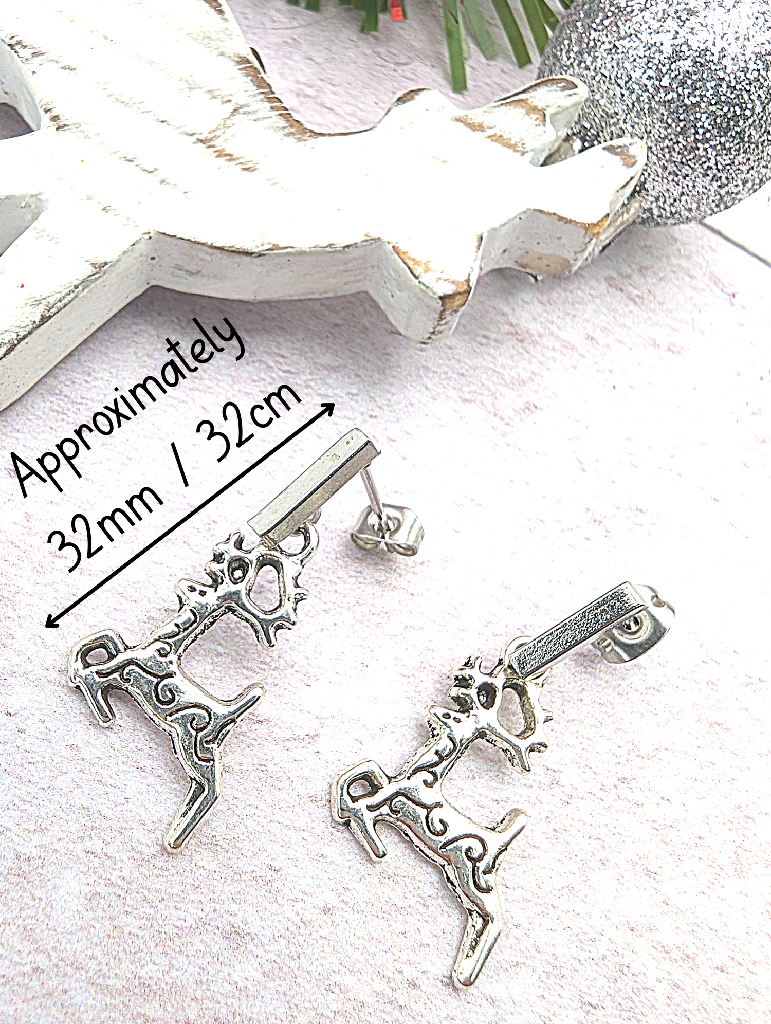 Displayed on a soft pink surface, two stainless steel reindeer charms gleam as part of the Prancing Reindeer Earrings by Silver Sunbird. In the background, a white Christmas tree decoration and a sparkling heart add to the festive scene. A label notes that these handmade earrings measure approximately 32mm.