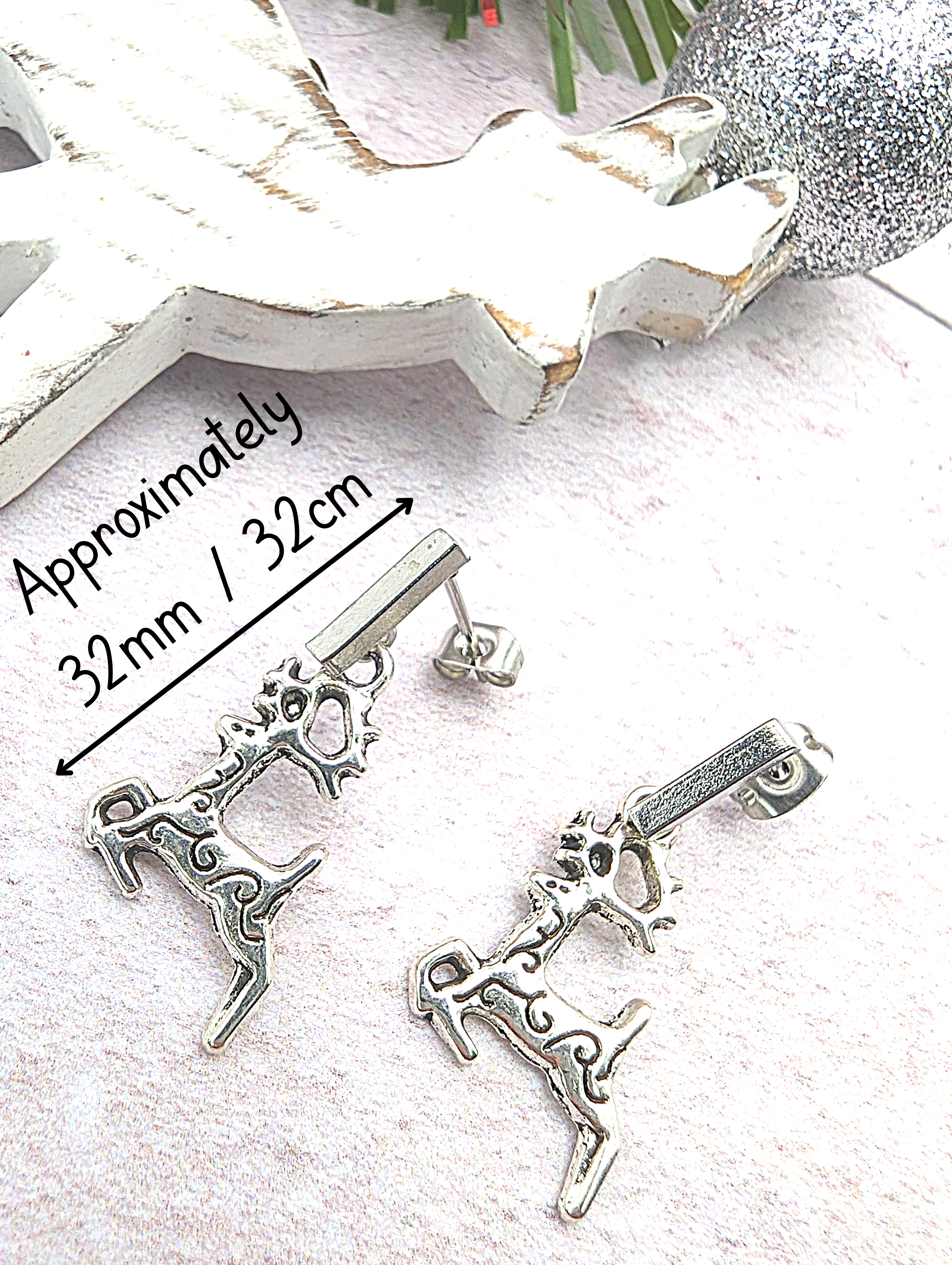 Displayed on a soft pink surface, two stainless steel reindeer charms gleam as part of the Prancing Reindeer Earrings by Silver Sunbird. In the background, a white Christmas tree decoration and a sparkling heart add to the festive scene. A label notes that these handmade earrings measure approximately 32mm.