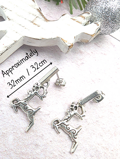 Displayed on a soft pink surface, two stainless steel reindeer charms gleam as part of the Prancing Reindeer Earrings by Silver Sunbird. In the background, a white Christmas tree decoration and a sparkling heart add to the festive scene. A label notes that these handmade earrings measure approximately 32mm.