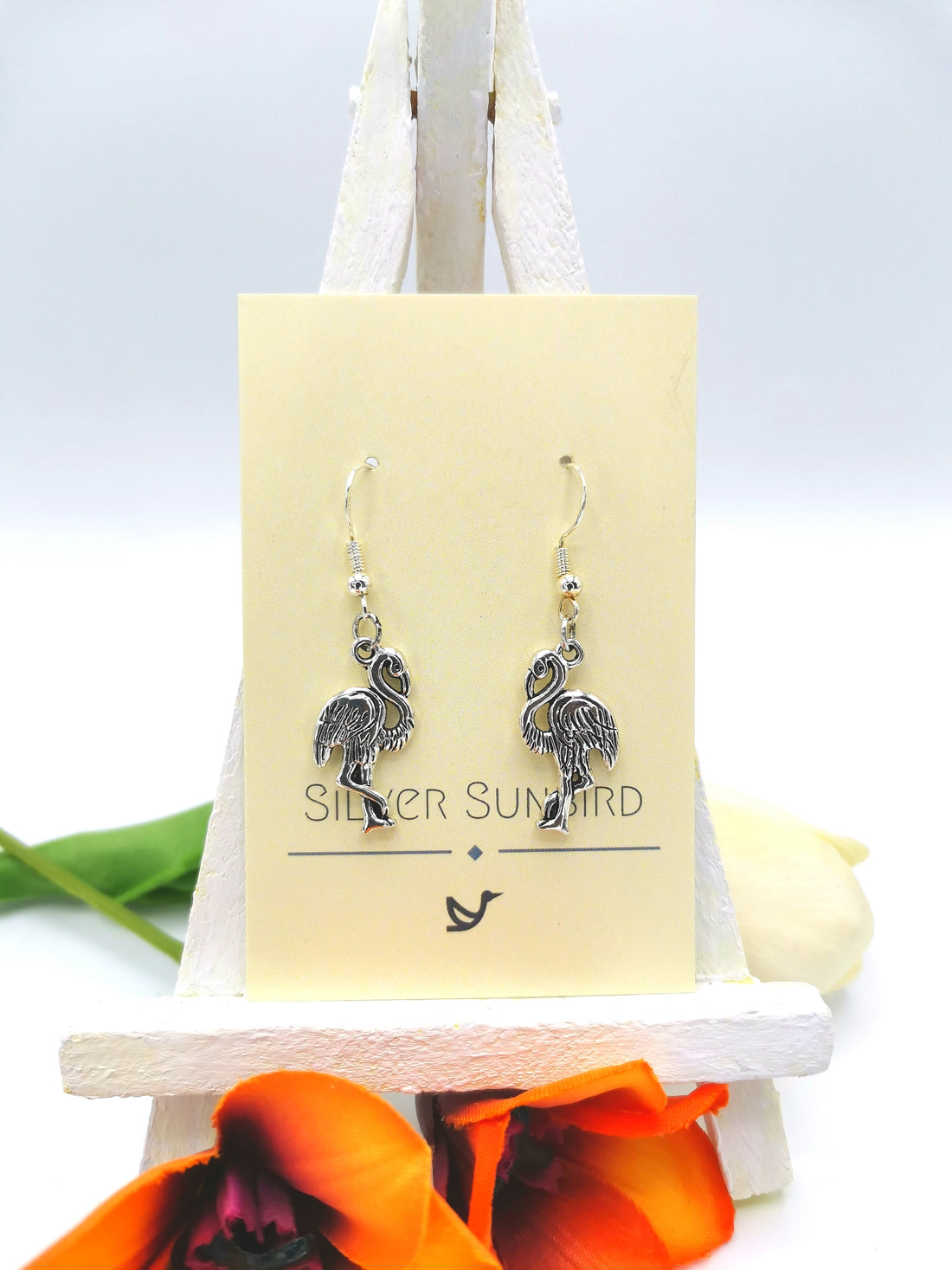 Close-up of a pair of Flocking Flamingo Earrings displayed on a yellow-coloured card with "Silver Sunbird" printed on it. The handmade jewellery is positioned on a small white easel, adorned with orange and white flowers at the base and some green leaves in the background.