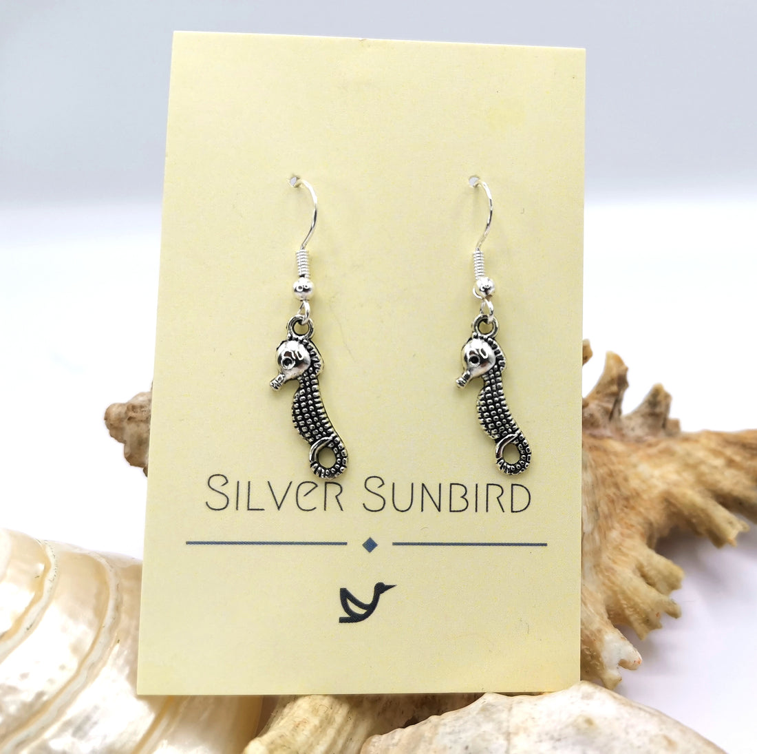 A pair of Spirited Seahorse Earrings by Silver Sunbird is elegantly showcased on a card with seashells in the background. Made from 925 sterling silver, these earrings boast intricate details and gracefully hang from silver hooks.