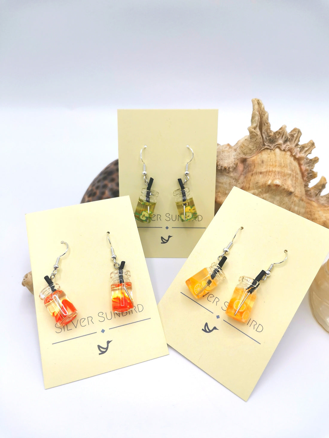 Three pairs of Party Cocktail Earrings by Silver Sunbird are shown on cream cards with a decorative seashell backdrop. Made of sterling silver, these handmade earrings come in vibrant green, orange, and yellow hues to add a splash of summer to any outfit.