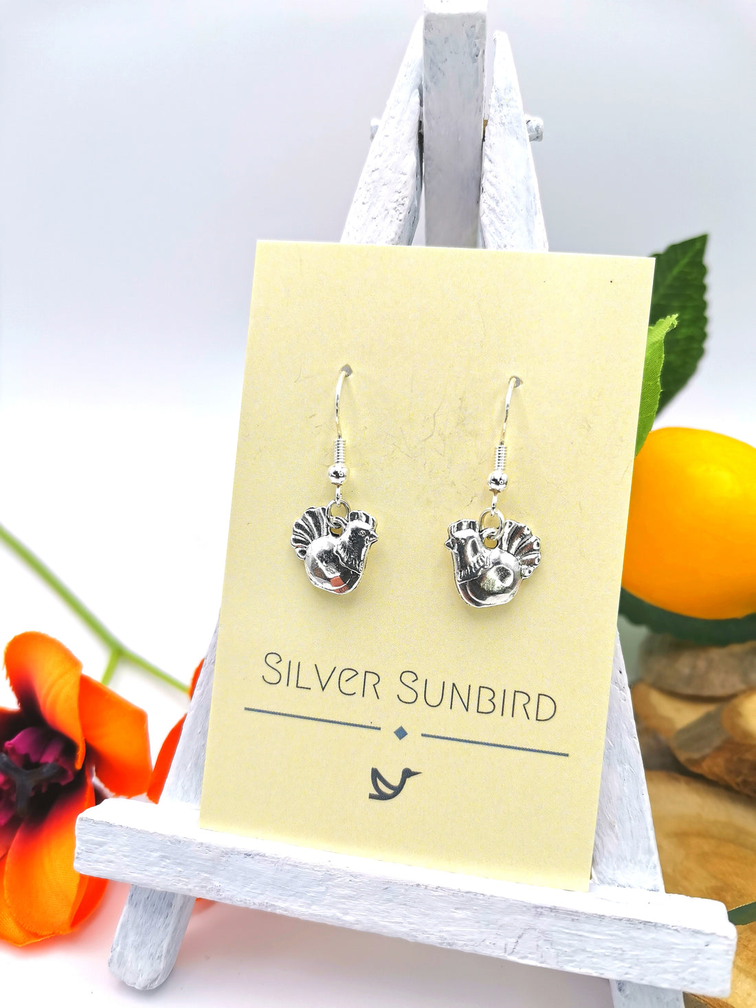The Charismatic Chicken Earrings by Silver Sunbird are showcased on an easel with a beige card. A bright orange flower and a yellow fruit in the background enhance their handmade charm.