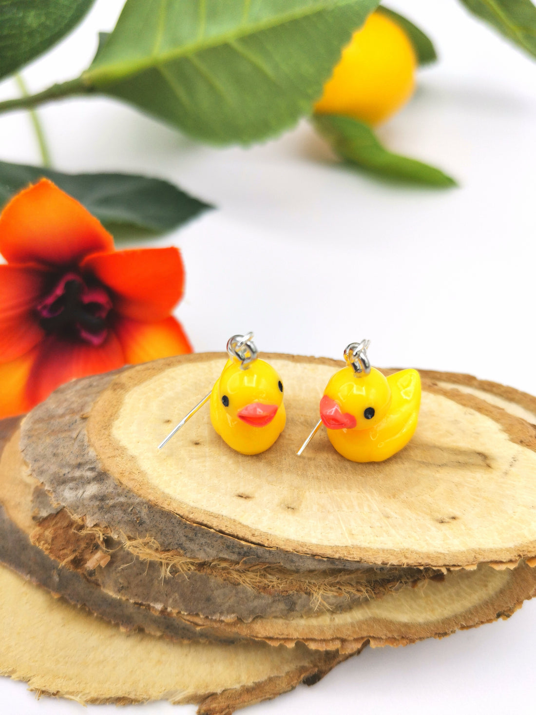 The Silver Sunbird Young Hearted Yellow Ducks Earrings feature two playful yellow duck charms on silver hooks, displayed on wooden slices among green leaves, an orange fruit, and an orange-red flower.