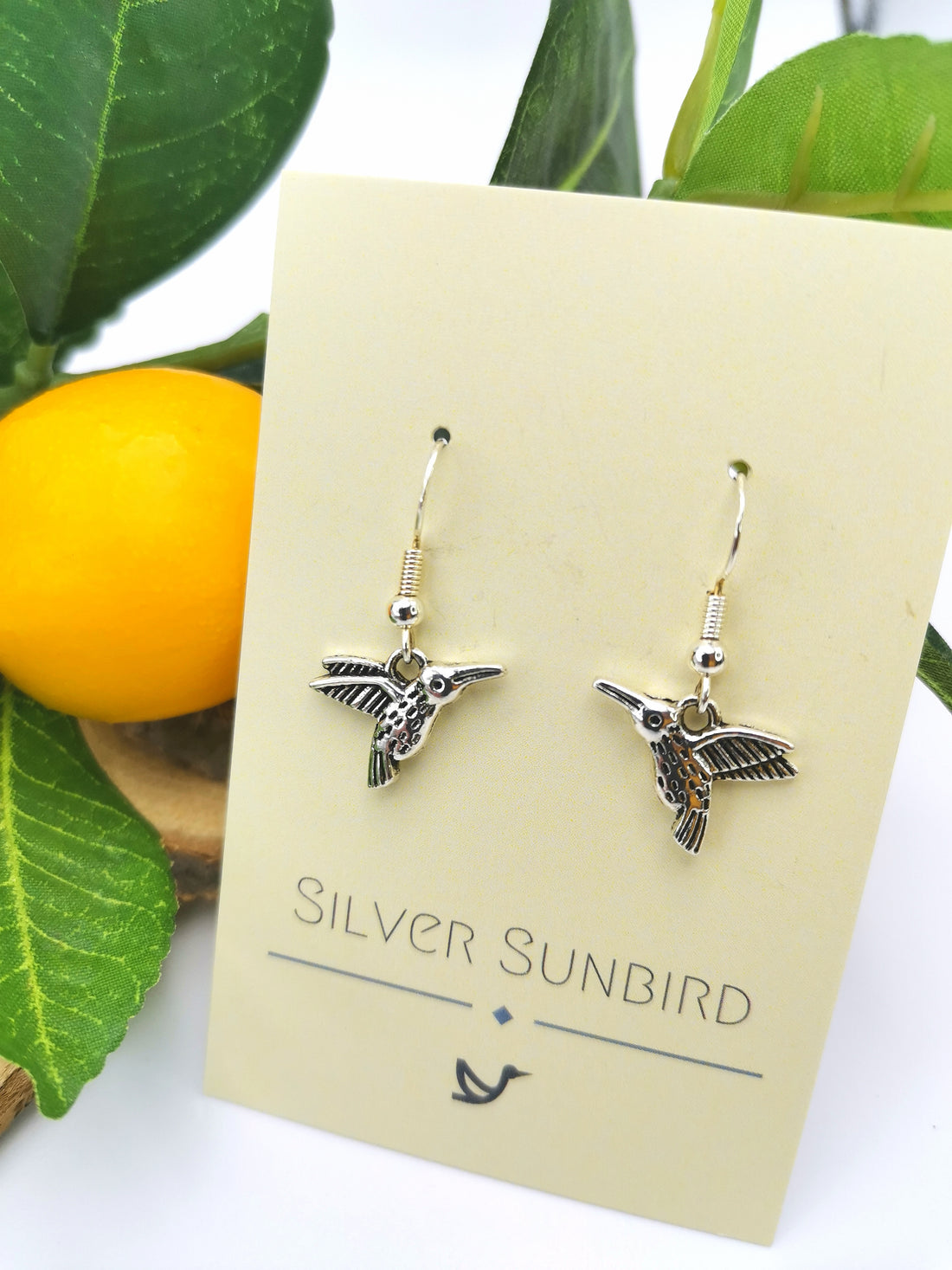 A pair of Heartfelt Hummingbird Earrings in sterling silver displayed on a cream-colored card with "Silver Sunbird" written in elegant letters. The background features green leaves and an orange fruit, enhancing the natural theme of this exquisite handmade jewellery.