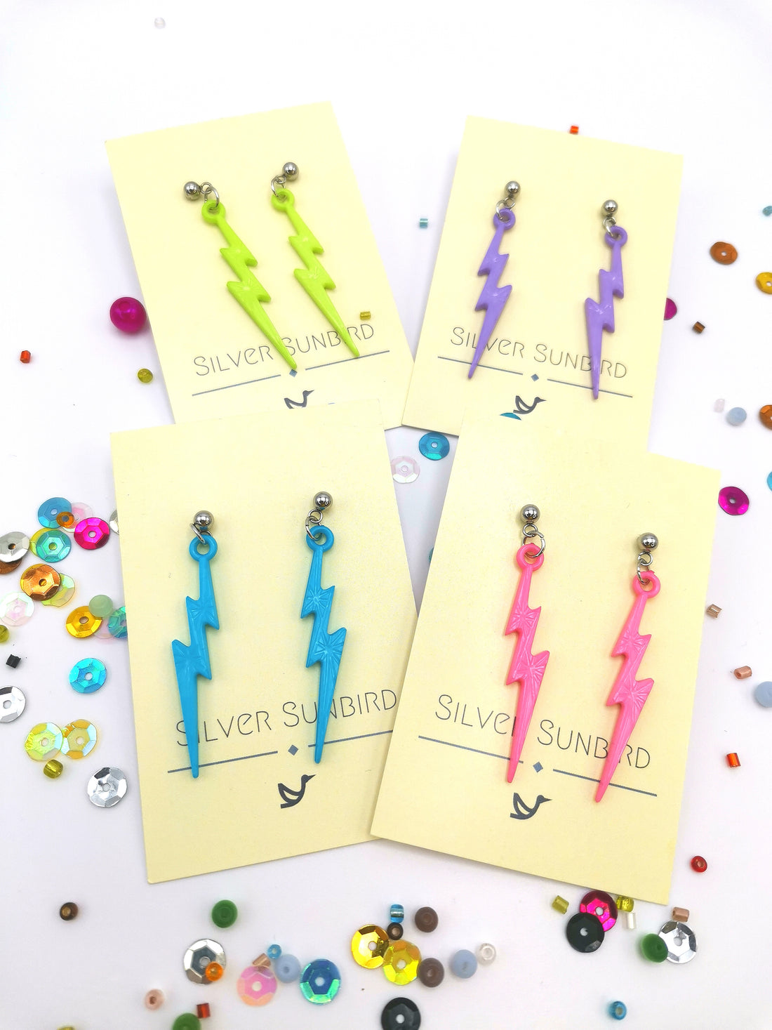 Five pairs of hypoallergenic stainless steel lightning bolt earrings in pastel hues—with vibrant shades like yellow, purple, blue, and pink—are displayed on "Silver Sunbird" branded cards. Sparkling sequins are artfully scattered around these striking accessories.