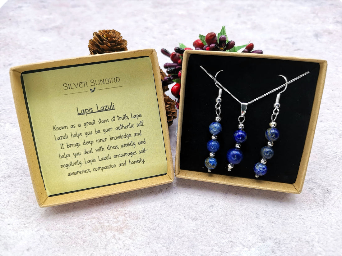 A stunning Lapis Lazuli Gemstone Jewellery Set by Silver Sunbird lies in an open box. The 925 sterling silver necklace and earrings showcase deep blue stones, accompanied by a card detailing their properties, with a decorative berry branch in the background.