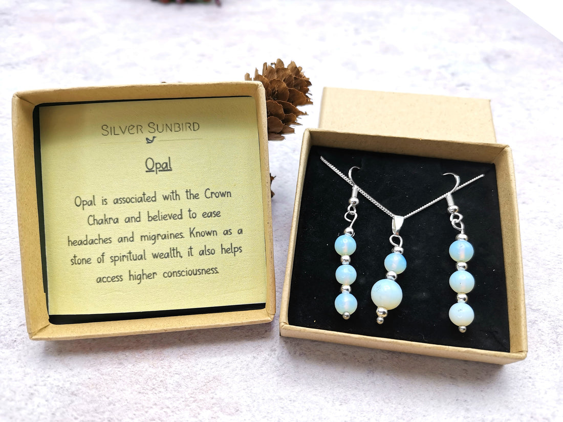 The Opal Gemstone Gift Set by Silver Sunbird is displayed in an open box on a light surface. This sterling silver set features a luminous opal necklace and matching earrings. An enclosed card details opal&