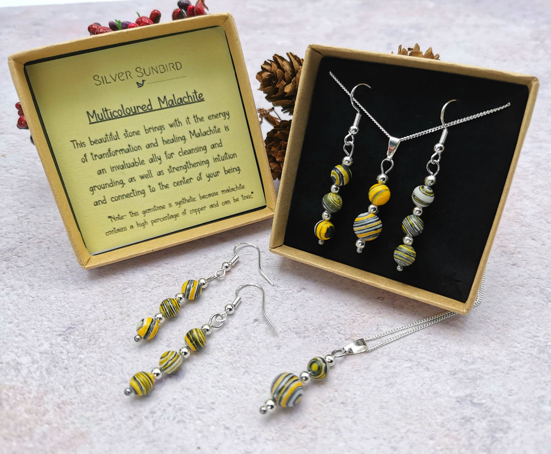 The Silver Sunbird Multi-coloured Malachite Gemstone Gift Set includes a necklace and earrings with multicolored malachite gemstones, presented in a "Multicoloured Malachite" box, accompanied by an energy card and a sprig of pine cones.
