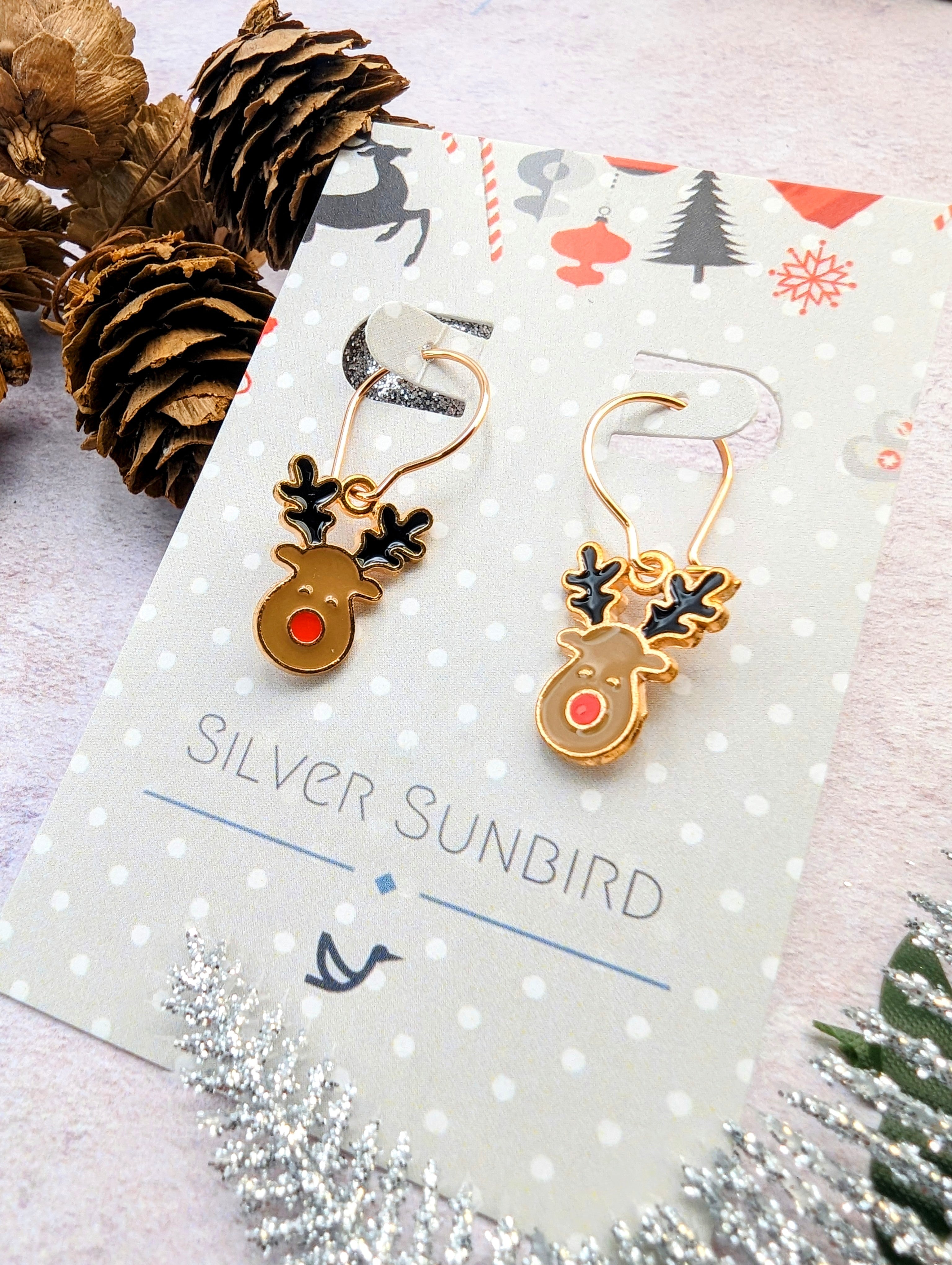 Rosie Reindeer Hoop Earrings by Silver Sunbird showcase gold reindeers with black antlers and red noses. They are presented on a festive, polka-dotted card adorned with pinecones and silver leaves.