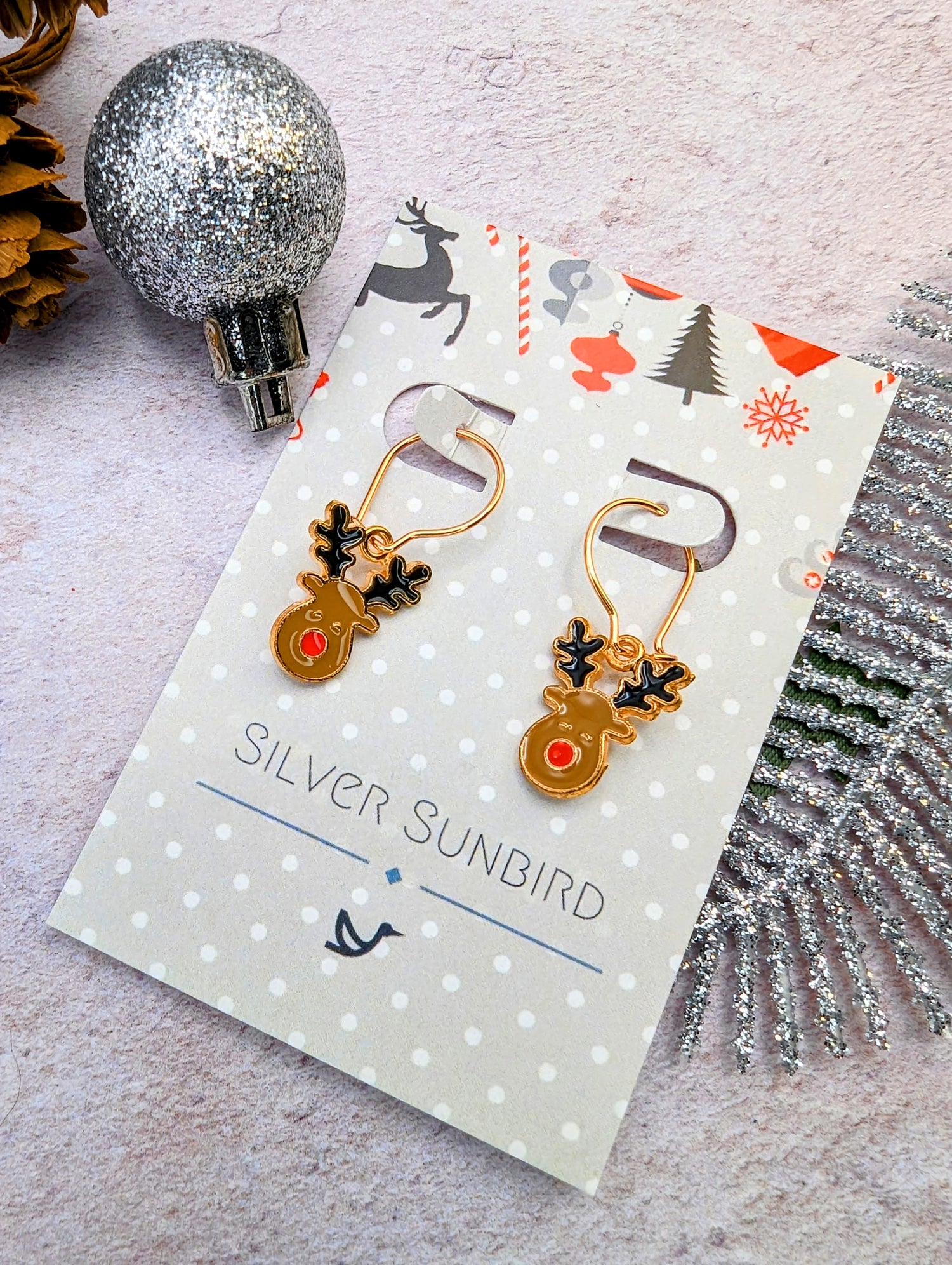 Rosie Reindeer Hoop Earrings from Silver Sunbird feature a festive design with red noses and black antlers, presented on a brand card. The background displays silver ornaments, pinecones, along with holly and reindeer motifs.