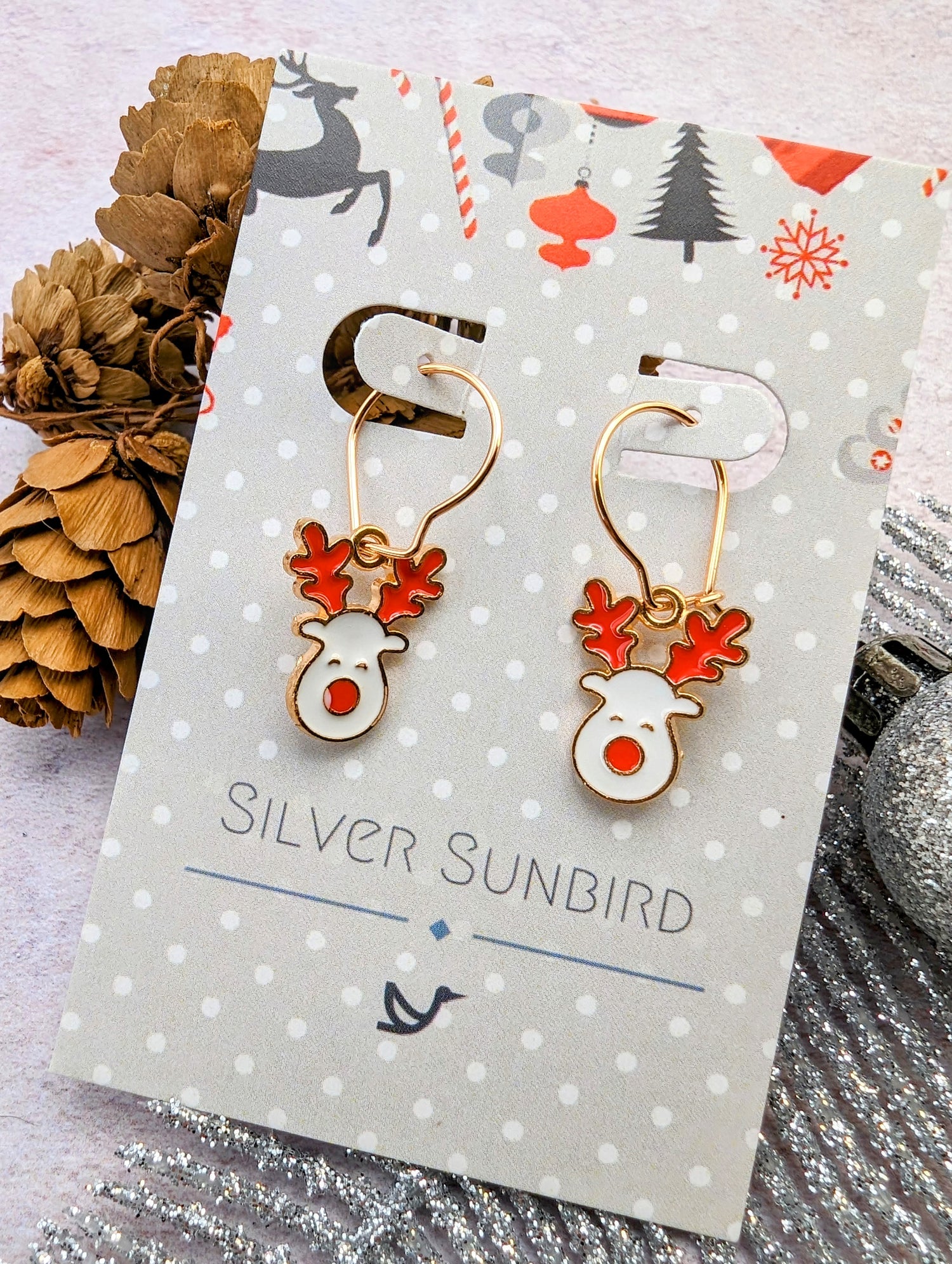 Displayed on a festive card from &quot;Silver Sunbird,&quot; the Rosie Reindeer Hoop Earrings feature charming reindeer heads with red antlers and noses. The background, decorated with pinecones and holiday-themed elements, perfectly complements these delightful enamel earrings.