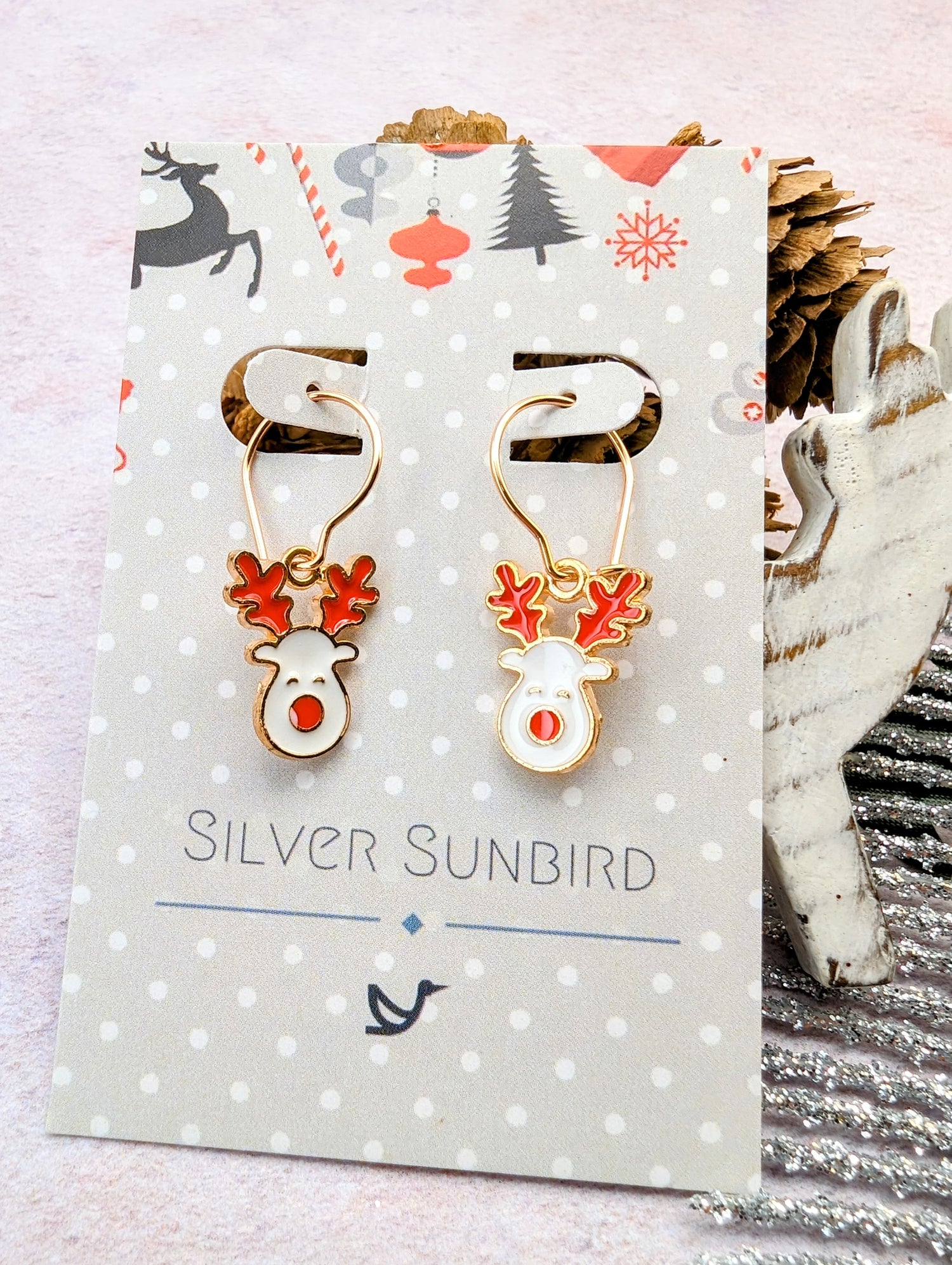 The Rosie Reindeer Hoop Earrings with red antlers and noses are elegantly showcased on a polka dot card branded &quot;Silver Sunbird.&quot; Featuring festive enamel designs, the background displays cheerful Christmas motifs and a charming pinecone decoration.