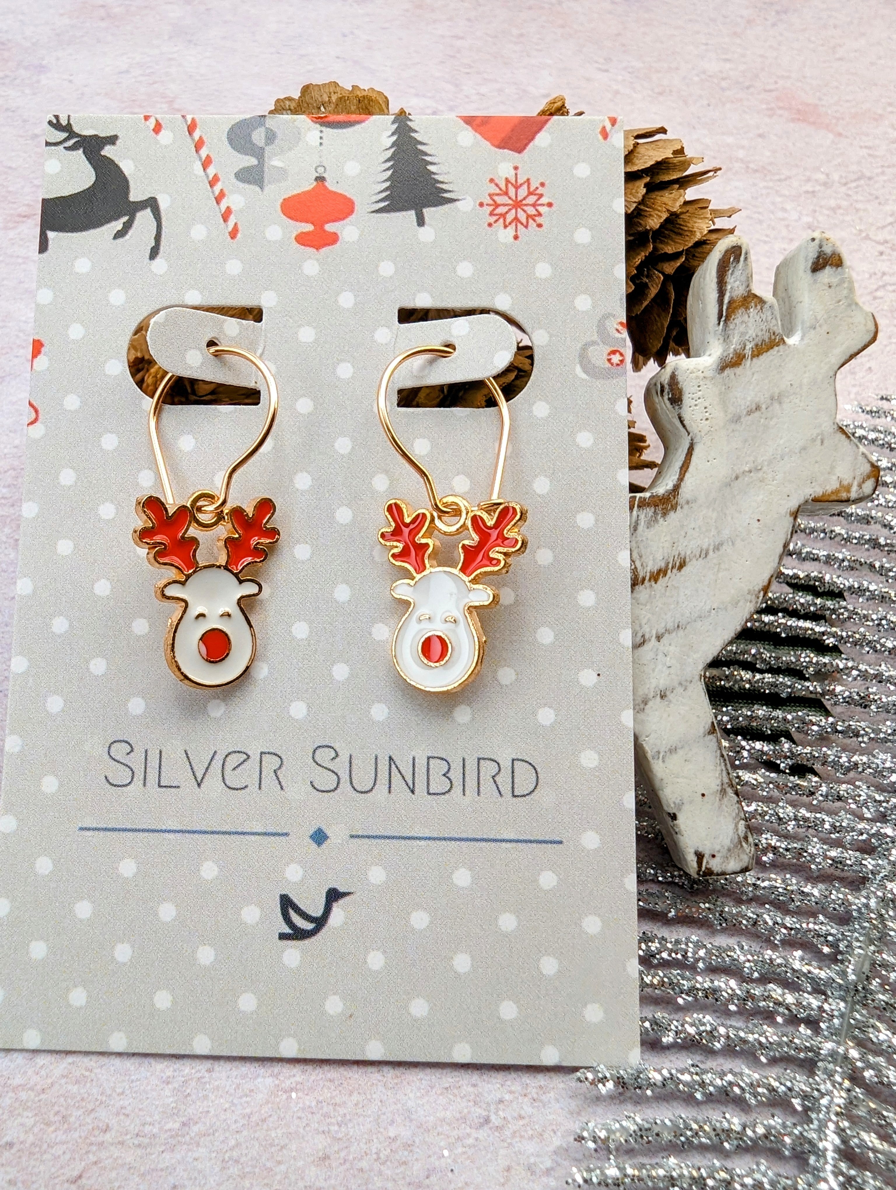 A pair of Rosie Reindeer Hoop Earrings, complete with red antlers and noses, are showcased on a card labeled &quot;Silver Sunbird.&quot; In the background, you can see pinecones, a wooden reindeer decoration, and a silver glittery garnish.