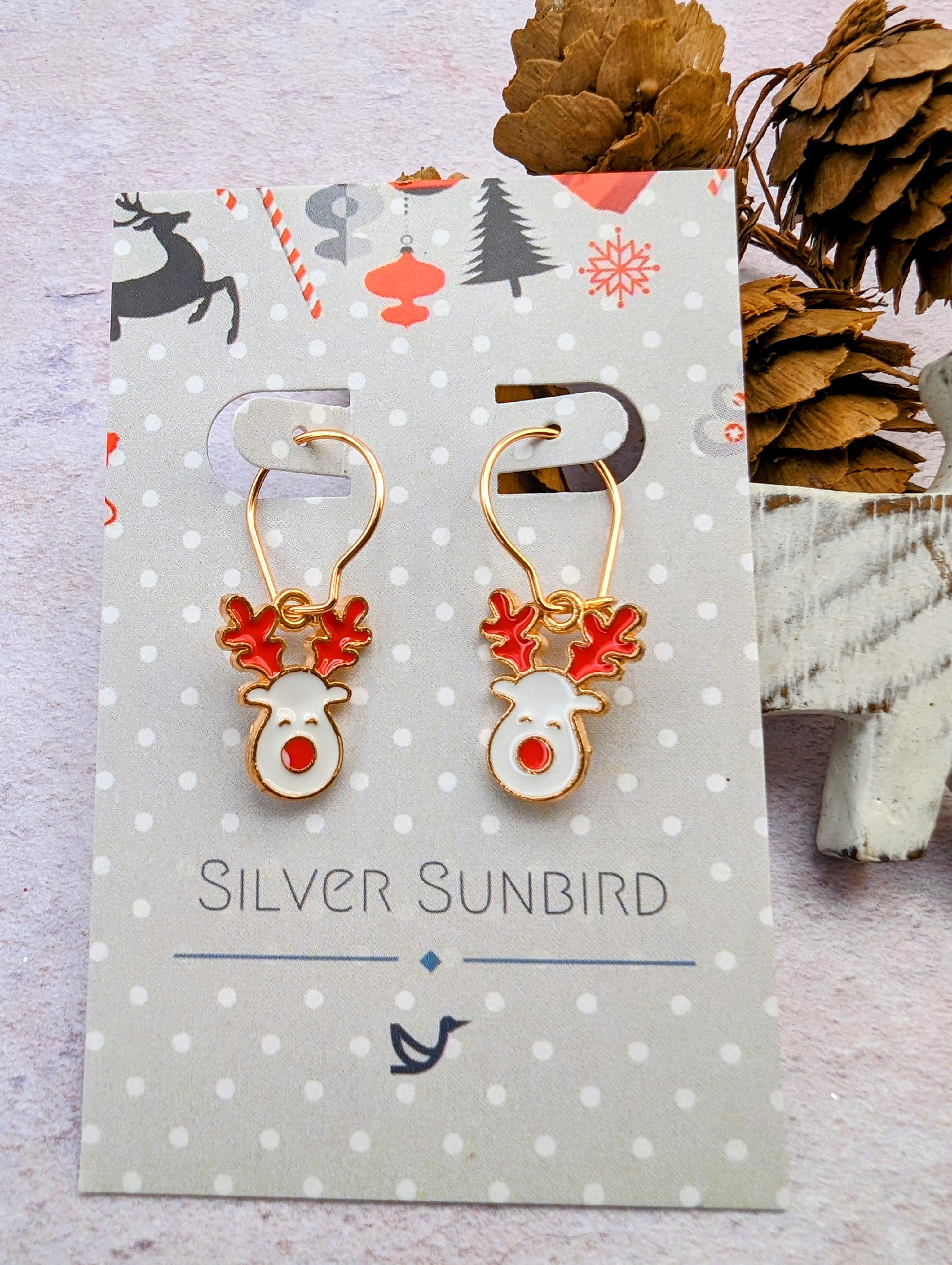 A pair of Rosie Reindeer Hoop Earrings by Silver Sunbird, showcasing festive enamel with red antlers and gold details, is elegantly displayed on a decorative card amidst small pine cones.