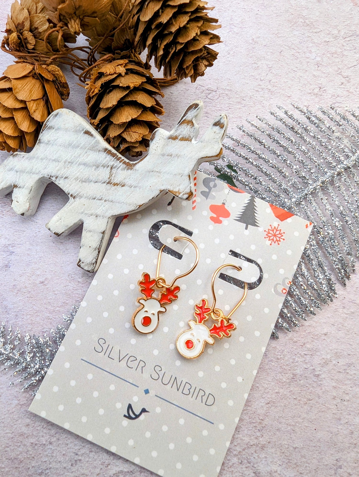 Adorned with red reindeer designs, the Rosie Reindeer Hoop Earrings by Silver Sunbird are elegantly presented on a branded card. Placed on a textured surface, they share space with pine cones, a wooden reindeer figurine, and a silver fern decoration, perfectly capturing the festive spirit.