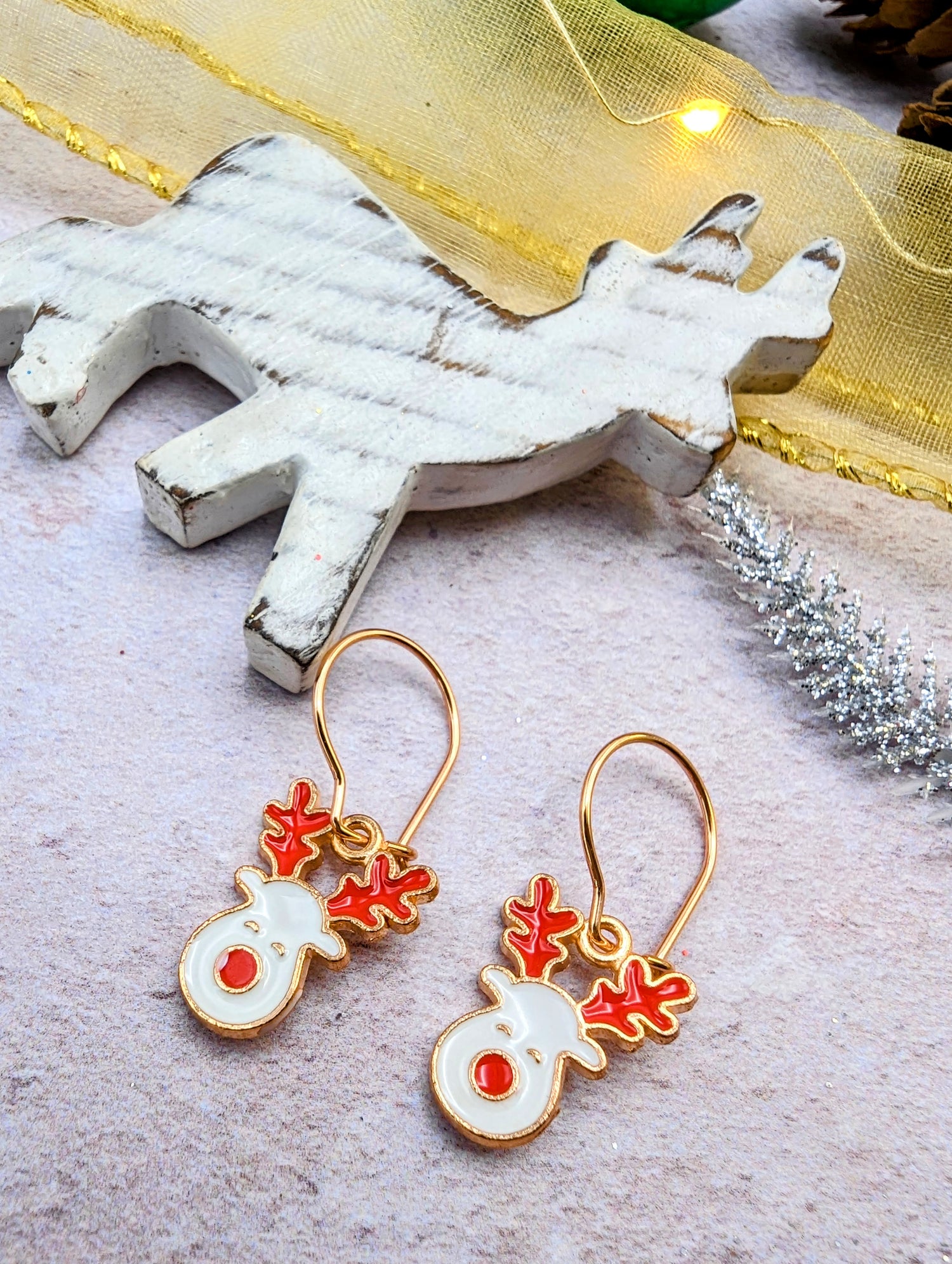The Rosie Reindeer Hoop Earrings by Silver Sunbird showcase charming enamel designs with red antlers and noses set against a light backdrop. In the background, a wooden reindeer decoration and silver branch are elegantly positioned.