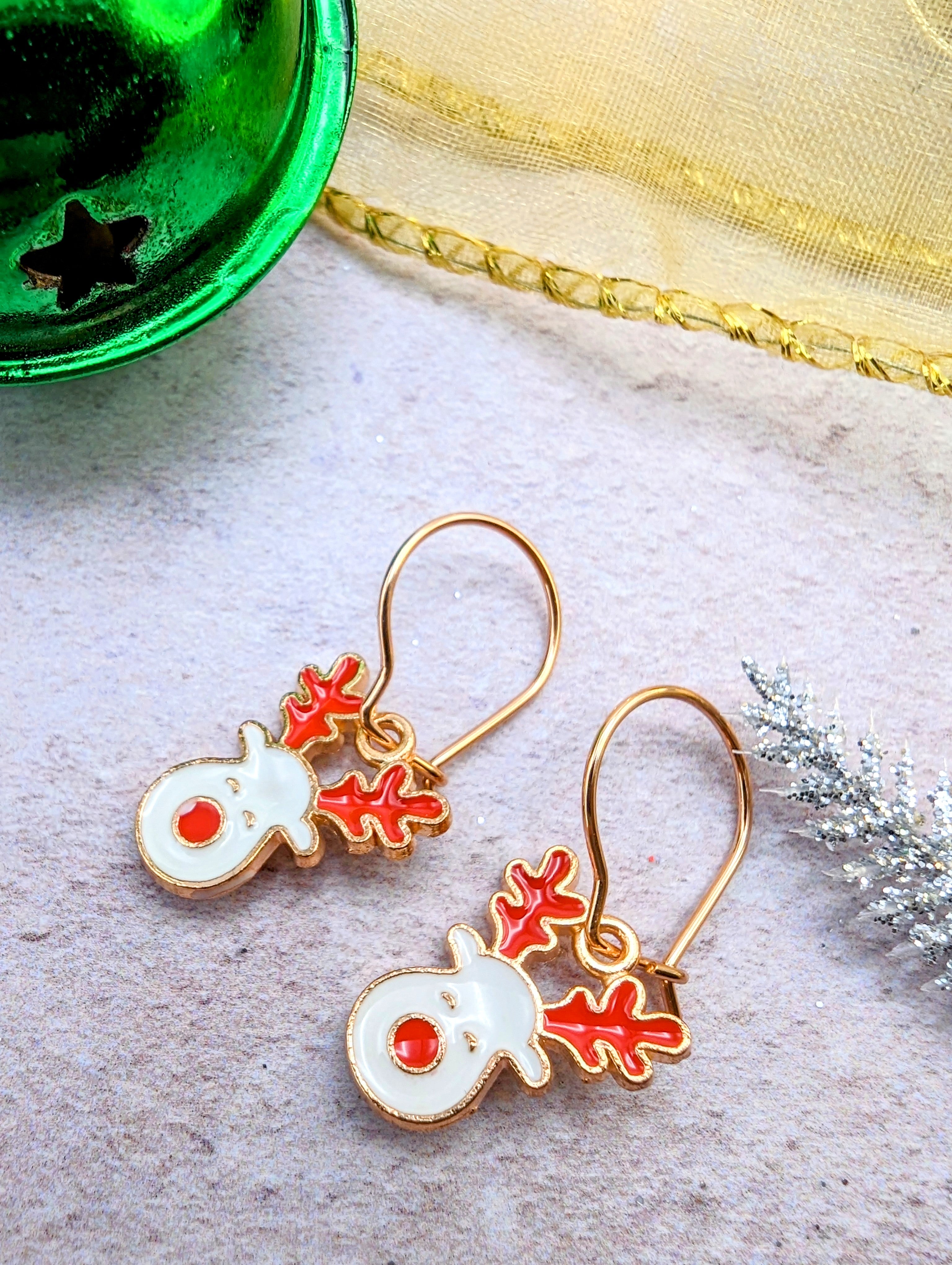 On a light gray surface, the Rosie Reindeer Hoop Earrings by Silver Sunbird, adorned with enamel reindeer heads complete with red noses and antlers, are elegantly displayed. Accompanying them in this delightful holiday-themed arrangement are a green bell and a silver branch, accentuating their playful charm.