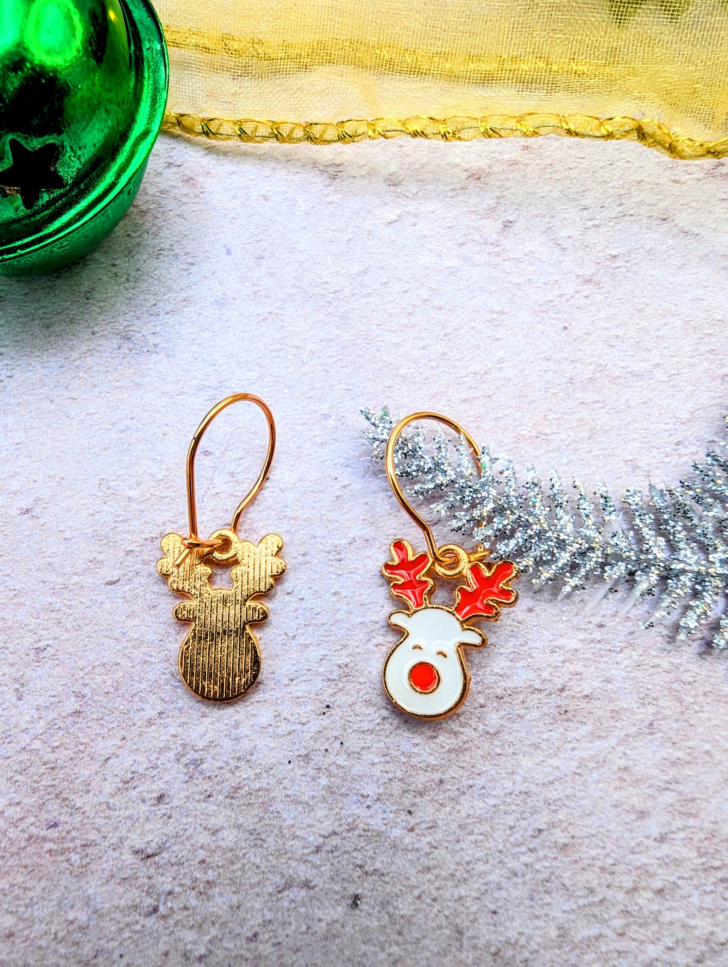 The Rosie Reindeer Hoop Earrings by Silver Sunbird showcase a reindeer-themed design on a textured surface. One earring highlights a simple gold silhouette, while the other displays an enamel white face with red antlers. A green ornament and silver pine branch in the background enhance the festive charm of these rose gold treasures.