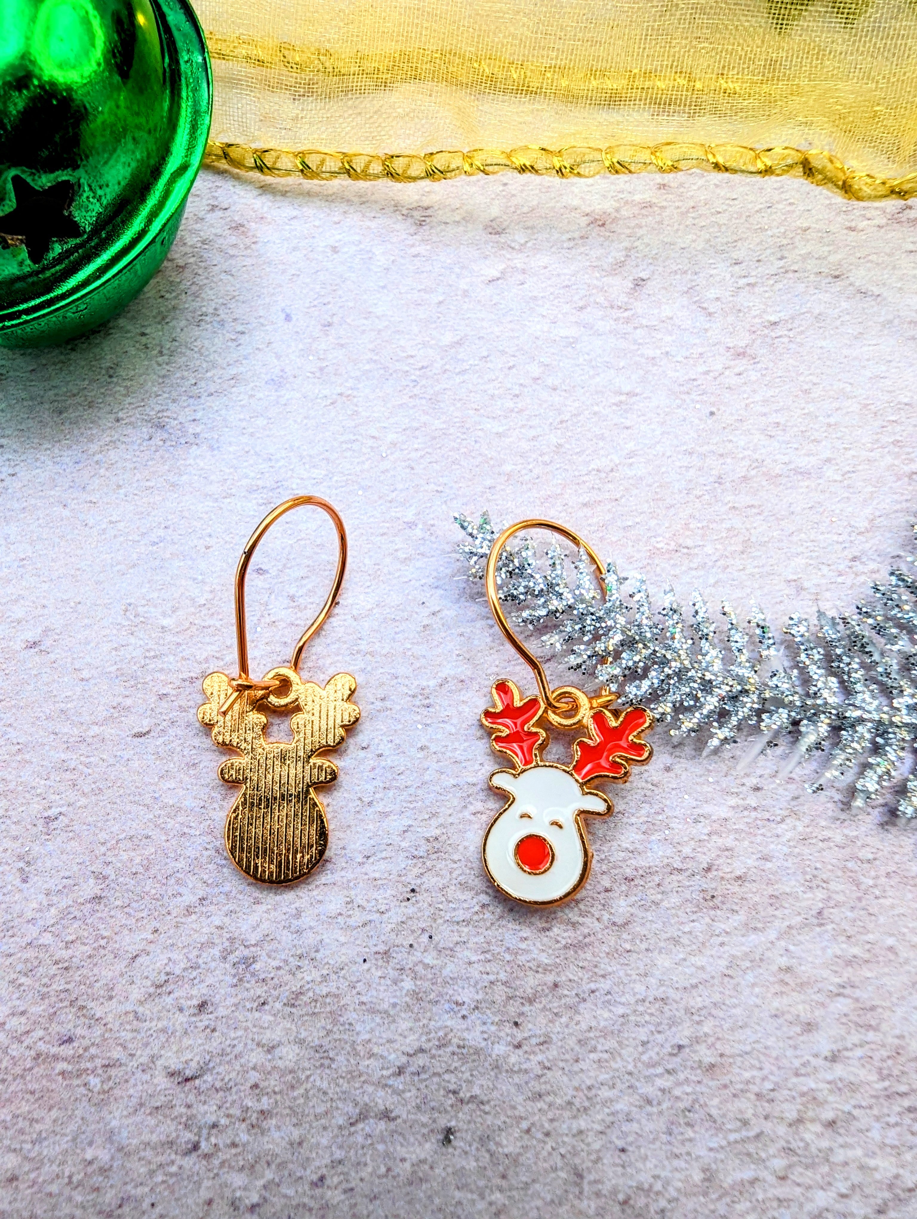 The Rosie Reindeer Hoop Earrings by Silver Sunbird showcase a reindeer-themed design on a textured surface. One earring highlights a simple gold silhouette, while the other displays an enamel white face with red antlers. A green ornament and silver pine branch in the background enhance the festive charm of these rose gold treasures.